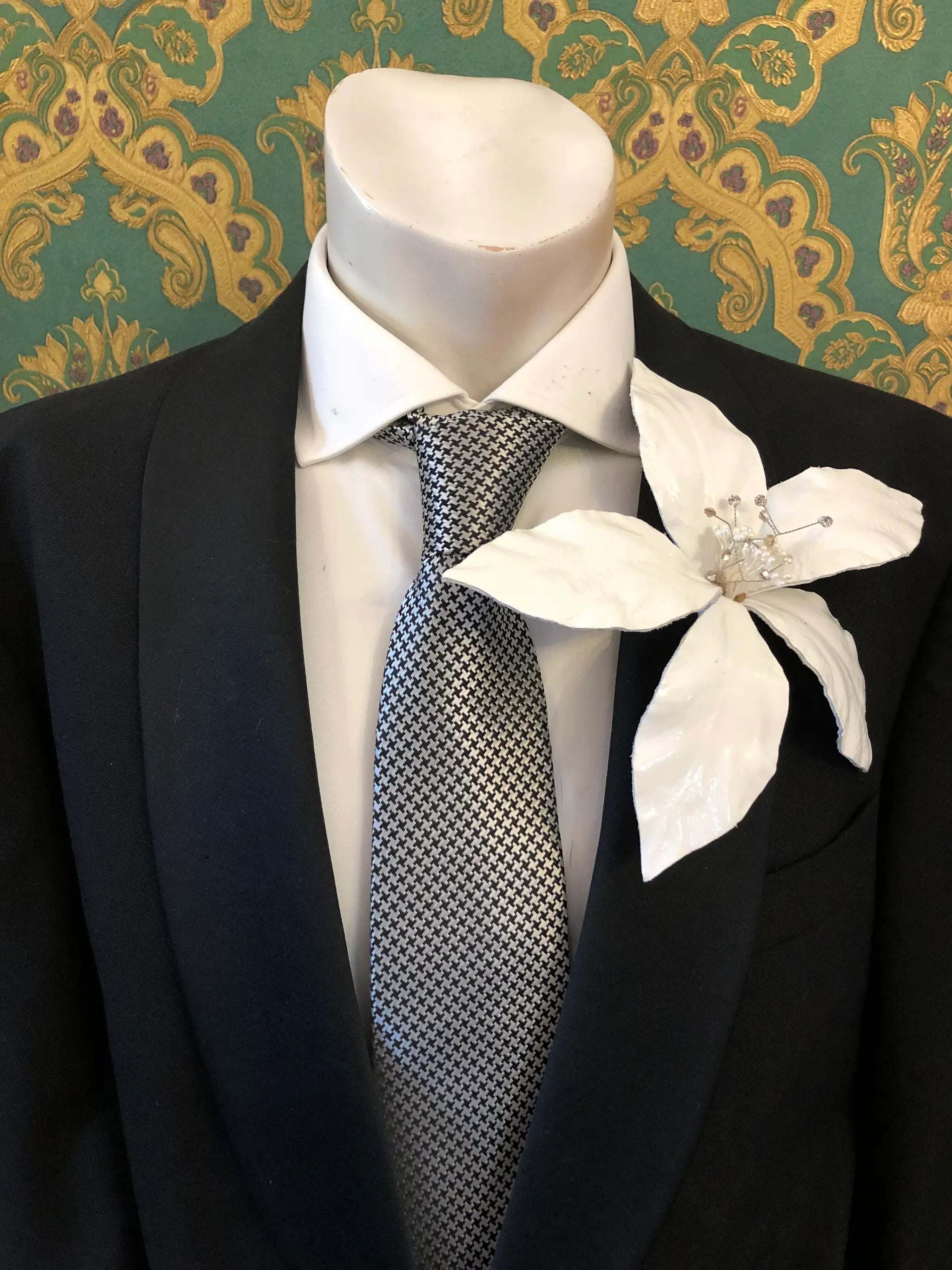 Broochella Men's Giglio patent leather flower brooch in white. It is pinned to a men's mannequin that is dressed in a black suit with a grey and silver houndstooth tie. The background is green and gold damask wallpaper. 