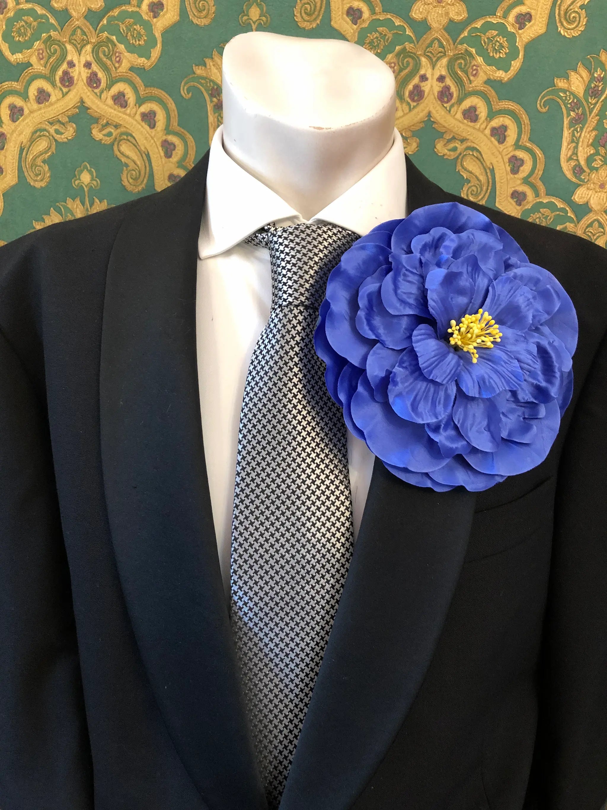 A large Broochella Men's Ibisco blue fabric flower brooch with a yellow centre,  pinned to the lapel of a black suit jacket over a white dress shirt and a black-and-white patterned tie.
