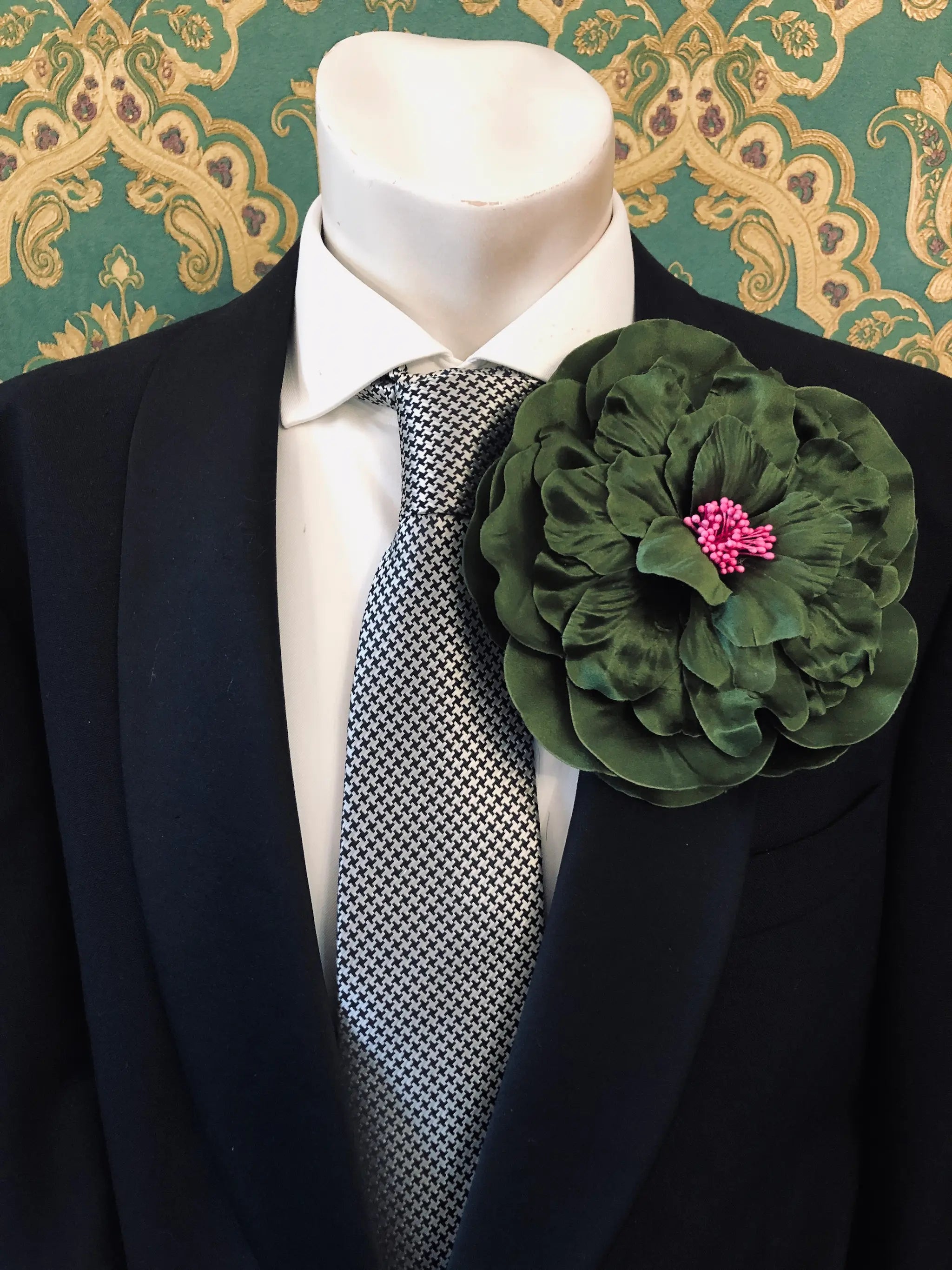 A large Broochella Men's Ibisco dark green fabric flower brooch with a bright pink centre,  pinned to the lapel of a black suit jacket over a white dress shirt and a black-and-white patterned tie.