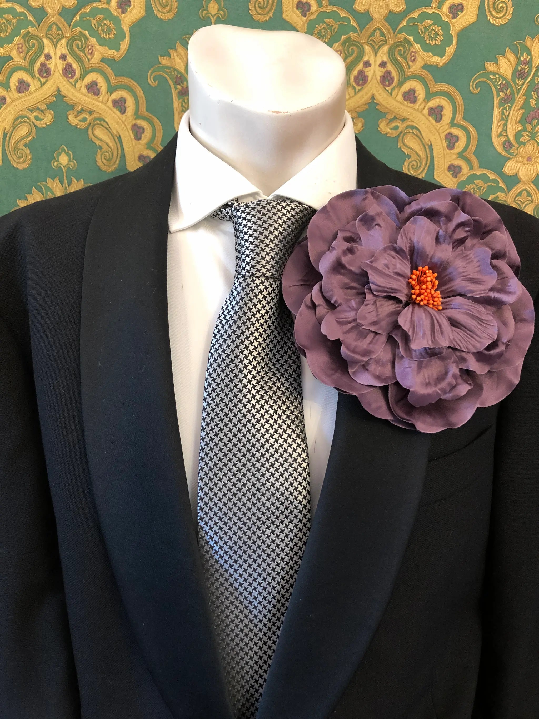 A large Broochella Men's Ibisco purple fabric flower brooch with an orange centre,  pinned to the lapel of a black suit jacket over a white dress shirt and a black-and-white patterned tie.