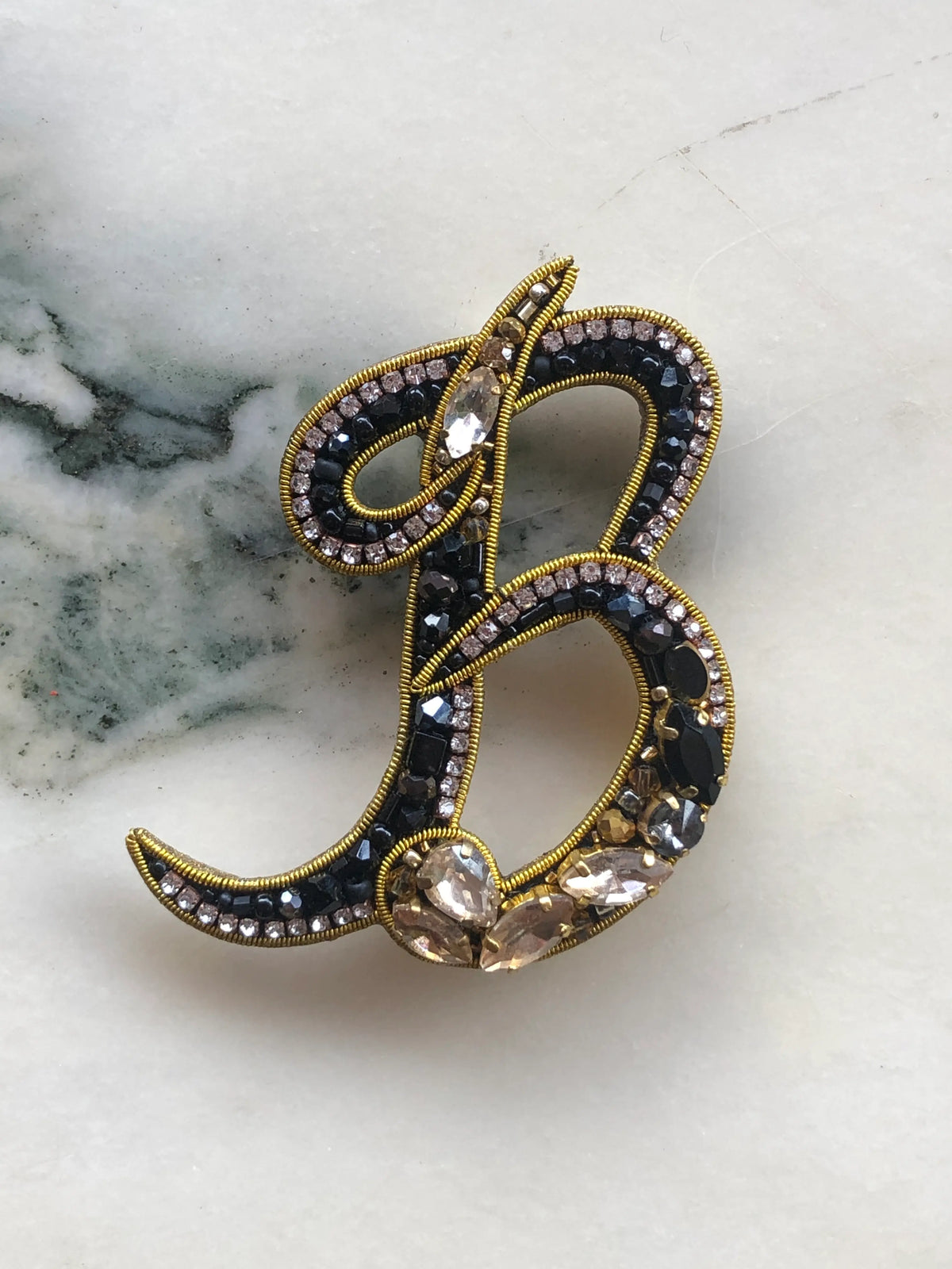 Broochella Men's Initial B Brooch embellished with black beads, clear crystals and champagne stones with gold detailing, photographed on a white marble background with soft grey veining.
