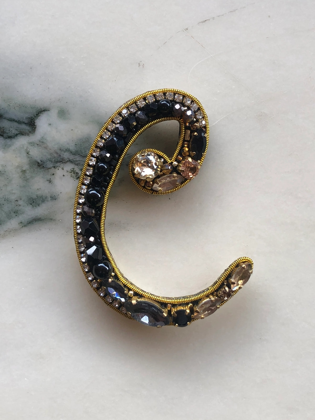 Broochella Men's Initial C Brooch embellished with black beads, clear crystals and champagne stones with gold detailing, photographed on a white marble background with soft grey veining.