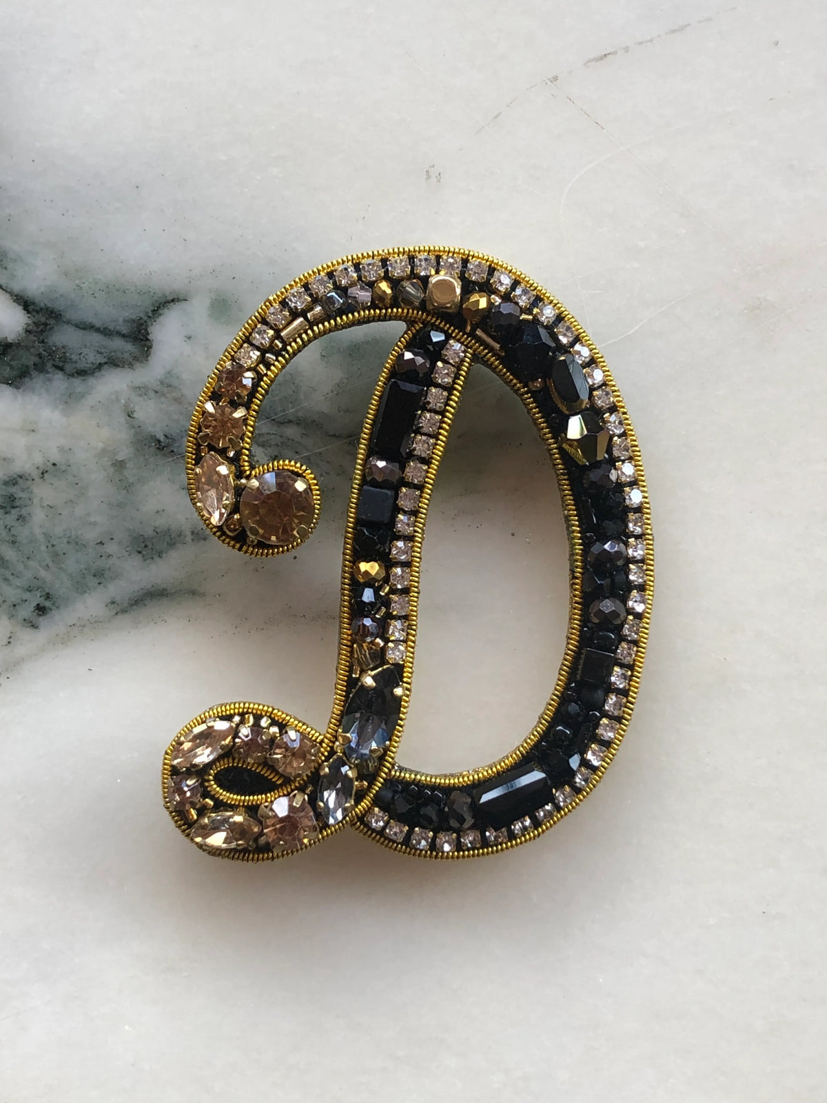 Broochella Men's Initial D Brooch embellished with black beads, clear crystals and champagne stones with gold detailing, photographed on a white marble background with soft grey veining.