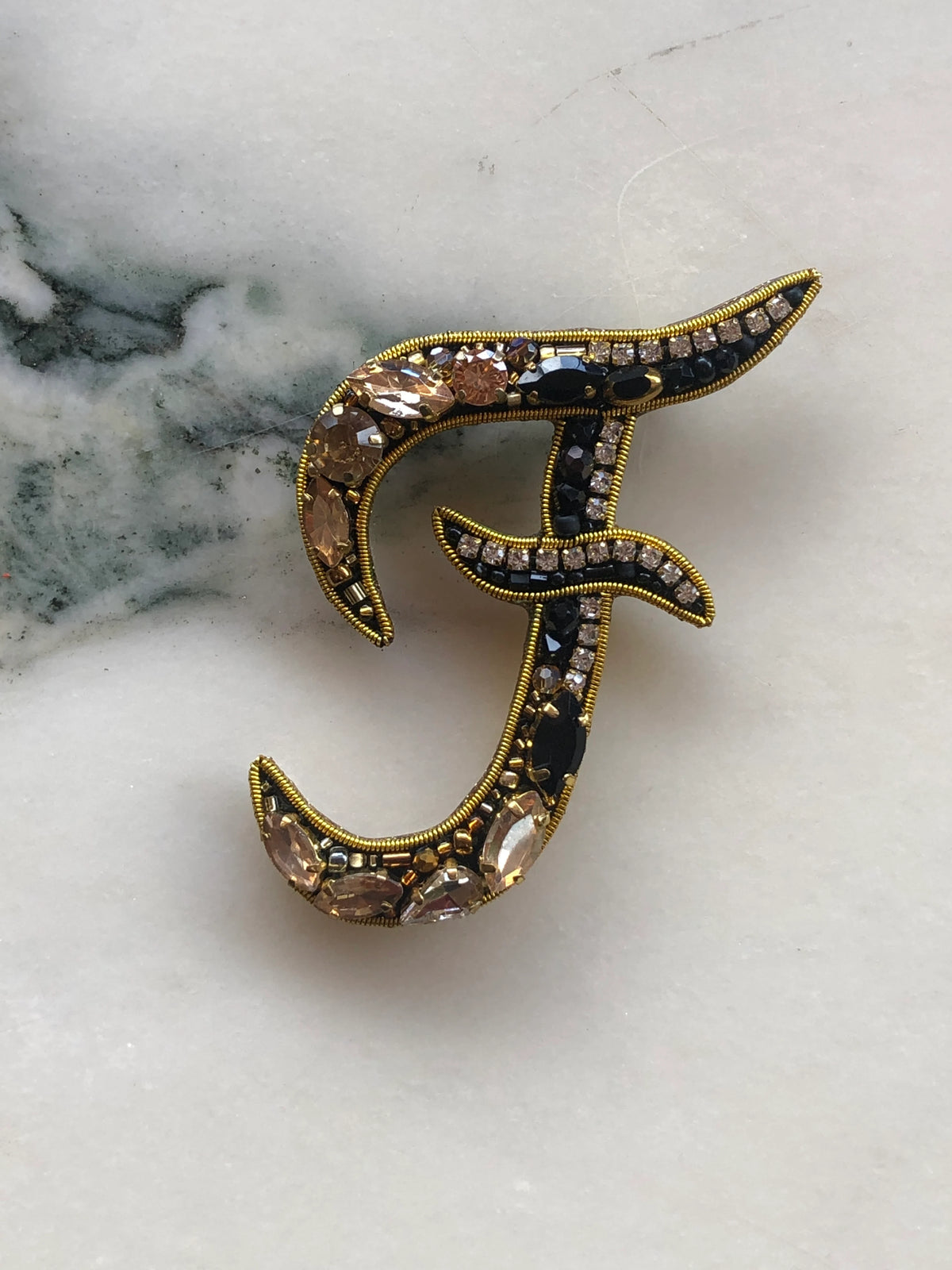 Broochella Men's Initial F Brooch embellished with black beads, clear crystals and champagne stones with gold detailing, photographed on a white marble background with soft grey veining.
