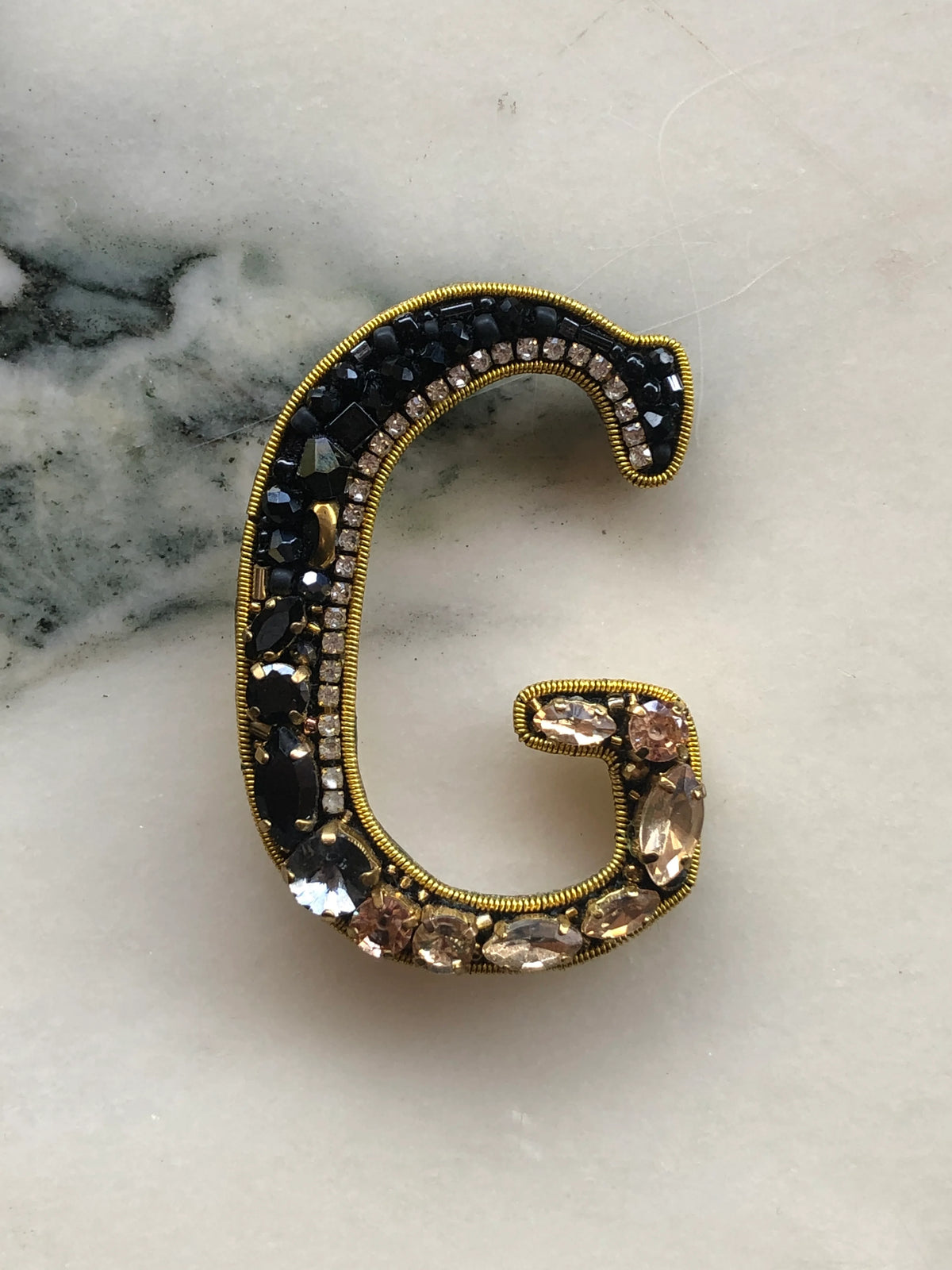Broochella Men's Initial G Brooch embellished with black beads, clear crystals and champagne stones with gold detailing, photographed on a white marble background with soft grey veining.