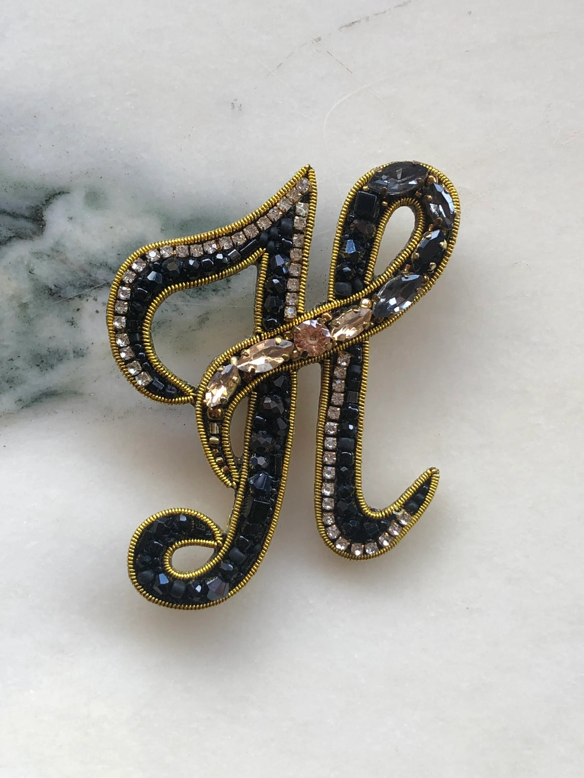 Broochella Men's Initial H Brooch embellished with black beads, clear crystals and champagne stones with gold detailing, photographed on a white marble background with soft grey veining.