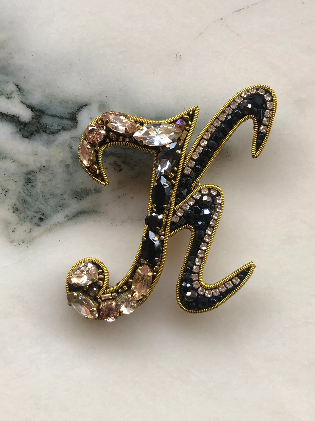 Broochella Men's Initial K Brooch embellished with black beads, clear crystals and champagne stones with gold detailing, photographed on a white marble background with soft grey veining.