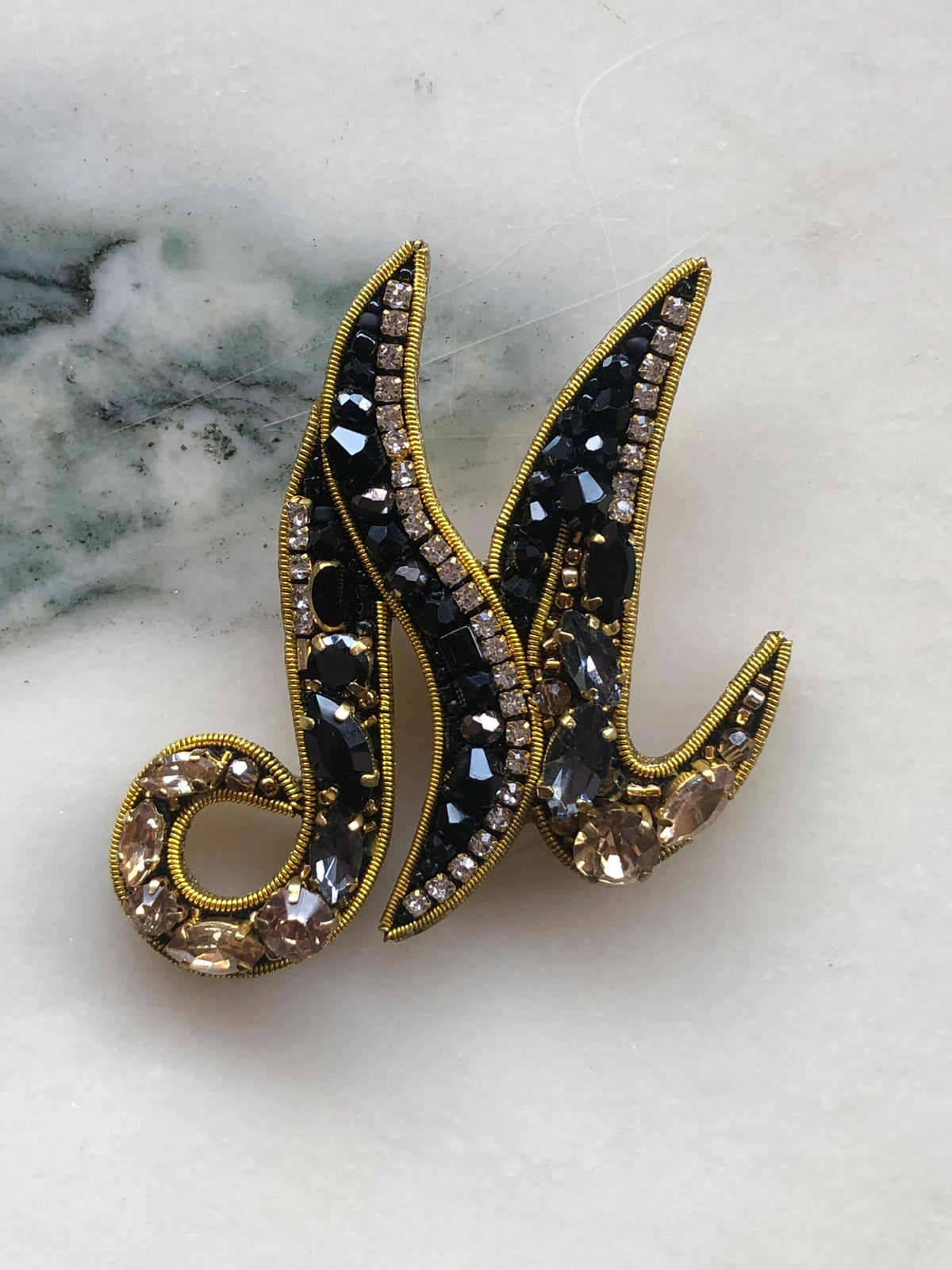 Broochella Men's Initial M Brooch embellished with black beads, clear crystals and champagne stones with gold detailing, photographed on a white marble background with soft grey veining.