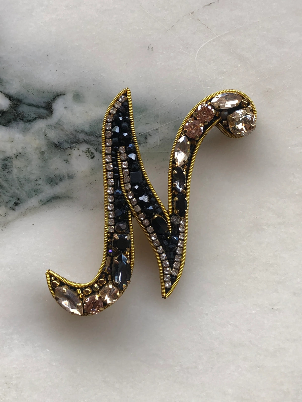 Broochella Men's Initial N Brooch embellished with black beads, clear crystals and champagne stones with gold detailing, photographed on a white marble background with soft grey veining.