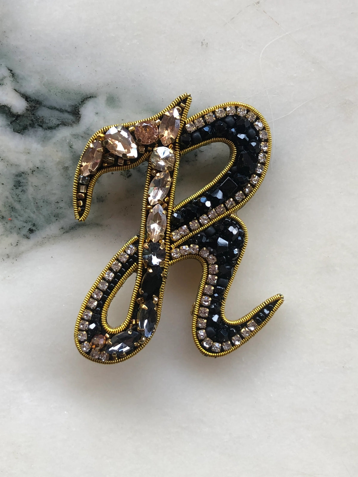 Broochella Men's Initial R Brooch embellished with black beads, clear crystals and champagne stones with gold detailing, photographed on a white marble background with soft grey veining.