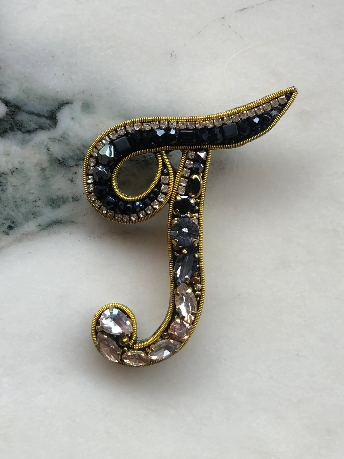 Broochella Men's Initial T Brooch embellished with black beads, clear crystals and champagne stones with gold detailing, photographed on a white marble background with soft grey veining.