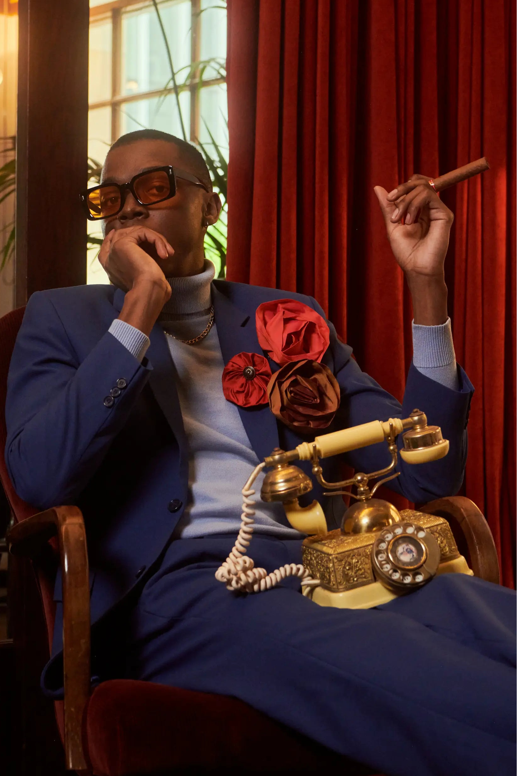 A man in a blue suit and sunglasses sits in a red armchair, holding a cigar. A vintage cream rotary telephone rests on his lap, and red curtains and warm interior lighting are visible in the background. He is wearing the Broochella men's lollipop brooch in red