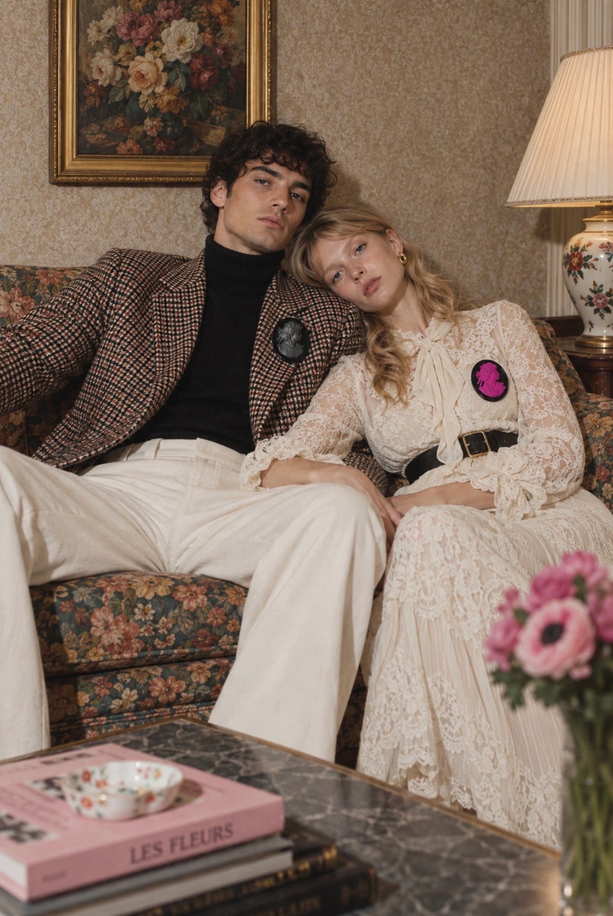 Broochella Men la Bella Figura cameo brooch. It is pictured on a male model in a tweed jacket and white trousers. He is sitting on a floral couch with a blonde model in a white lace dress. 