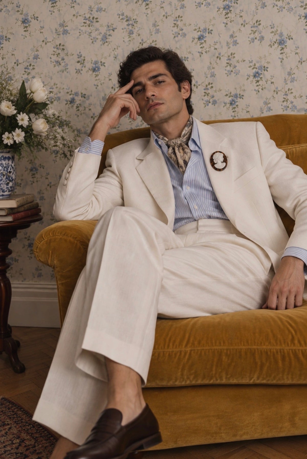 A man wearing a cream-colored three-piece suit with a light blue striped shirt and a patterned neck scarf, sitting on a mustard yellow velvet armchair in front of a blue floral wallpaper background. He is wearing a Broochella La Bella Figura Men's tortoiseshell cameo brooch.