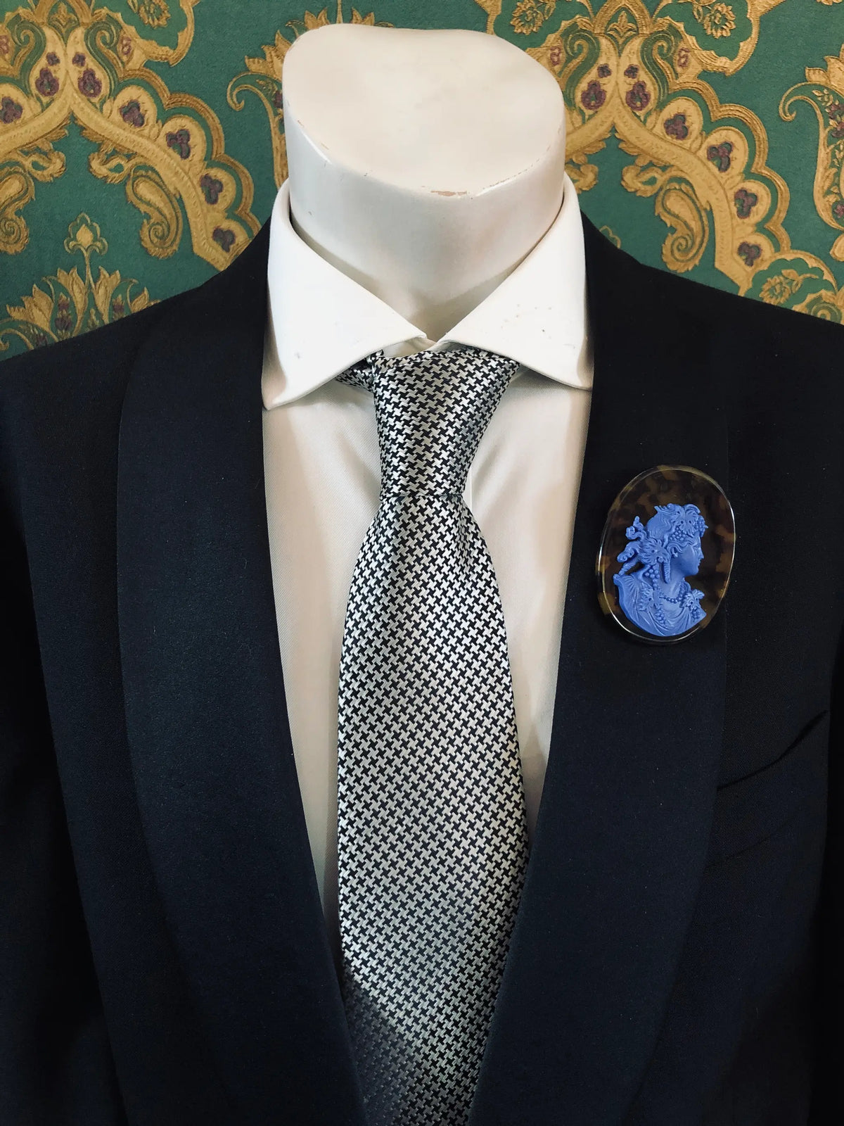 A black suit jacket on a mannequin torso, styled with a white dress shirt and a silver-and-black checkered tie. A Broochella Men's La Bella Figura bright blue and tortoiseshell cameo oval brooch is pinned to the left chest.
