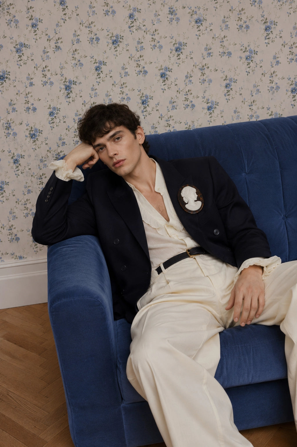 A man reclining on a blue velvet sofa against a floral wallpaper background, wearing a dark navy blazer with a Broochella Men's La Bella Figura tortoiseshell white cameo brooch cream open-collar shirt, cream trousers, and a black belt.
