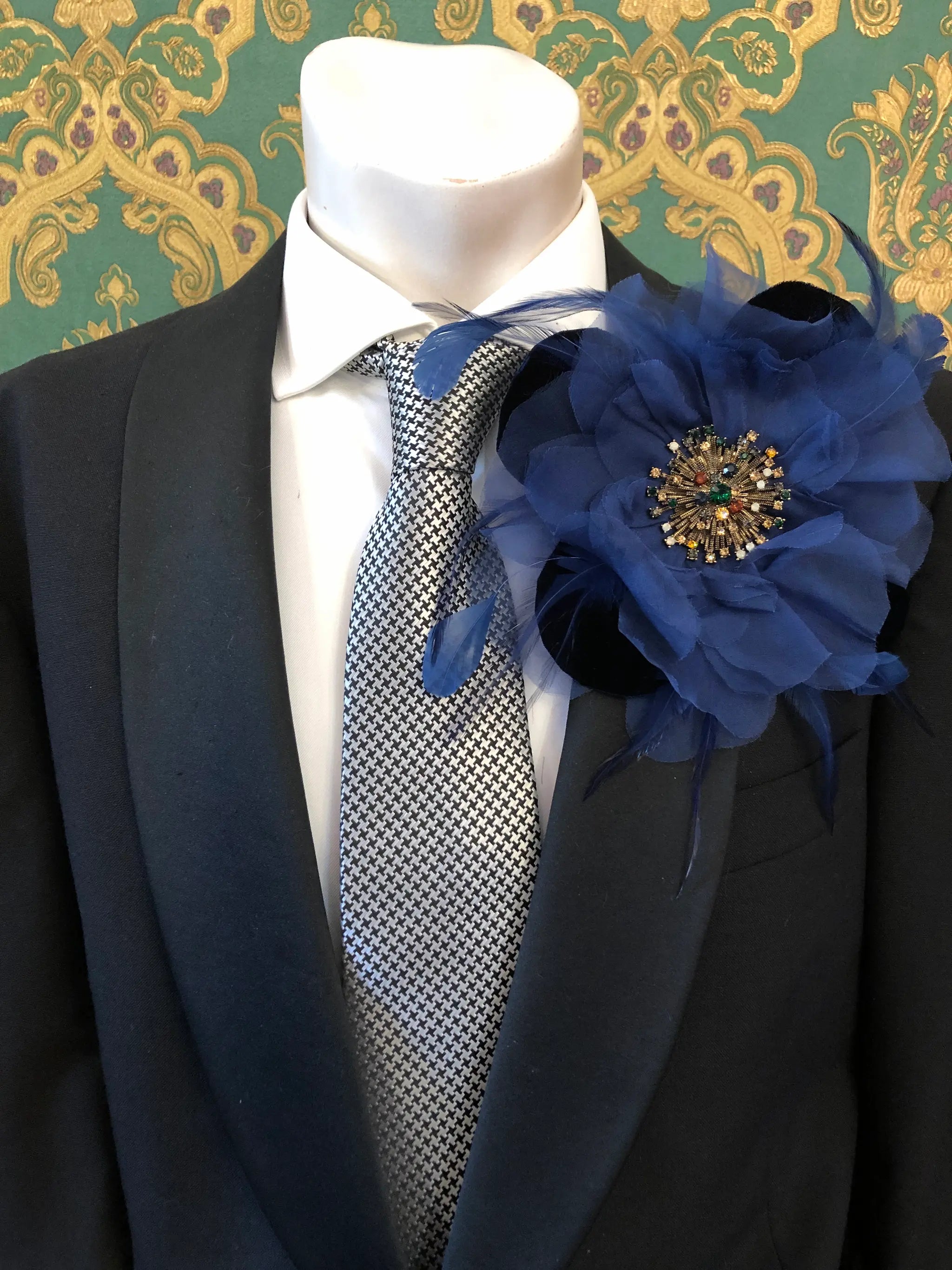 A black suit jacket on a mannequin torso, styled with a white dress shirt, a grey and white patterned tie, and a Broochella Men's la Duchessa large navy blue fabric flower brooch with a jewelled centre, set against a green damask wallpaper background.