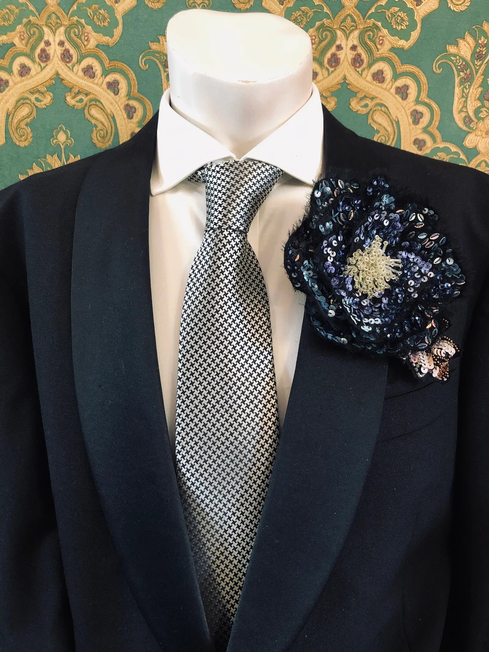 A close-up of a formal outfit on a mannequin torso, featuring a black suit jacket, white dress shirt, patterned grey and white tie, and a Broochella Men's Luminosa dark blue sequinned floral hand made flower brooch. The background is green and gold damask wallpaper. 