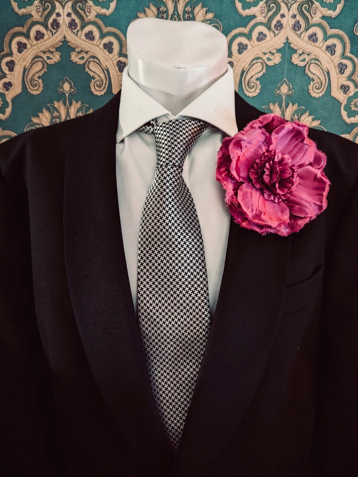 A men's formal outfit displayed on a white mannequin torso, featuring a black suit jacket, a white dress shirt, a black-and-white patterned tie, and a Broochella malva men's flower brooch on the lapel, set against ornate teal damask wallpaper.