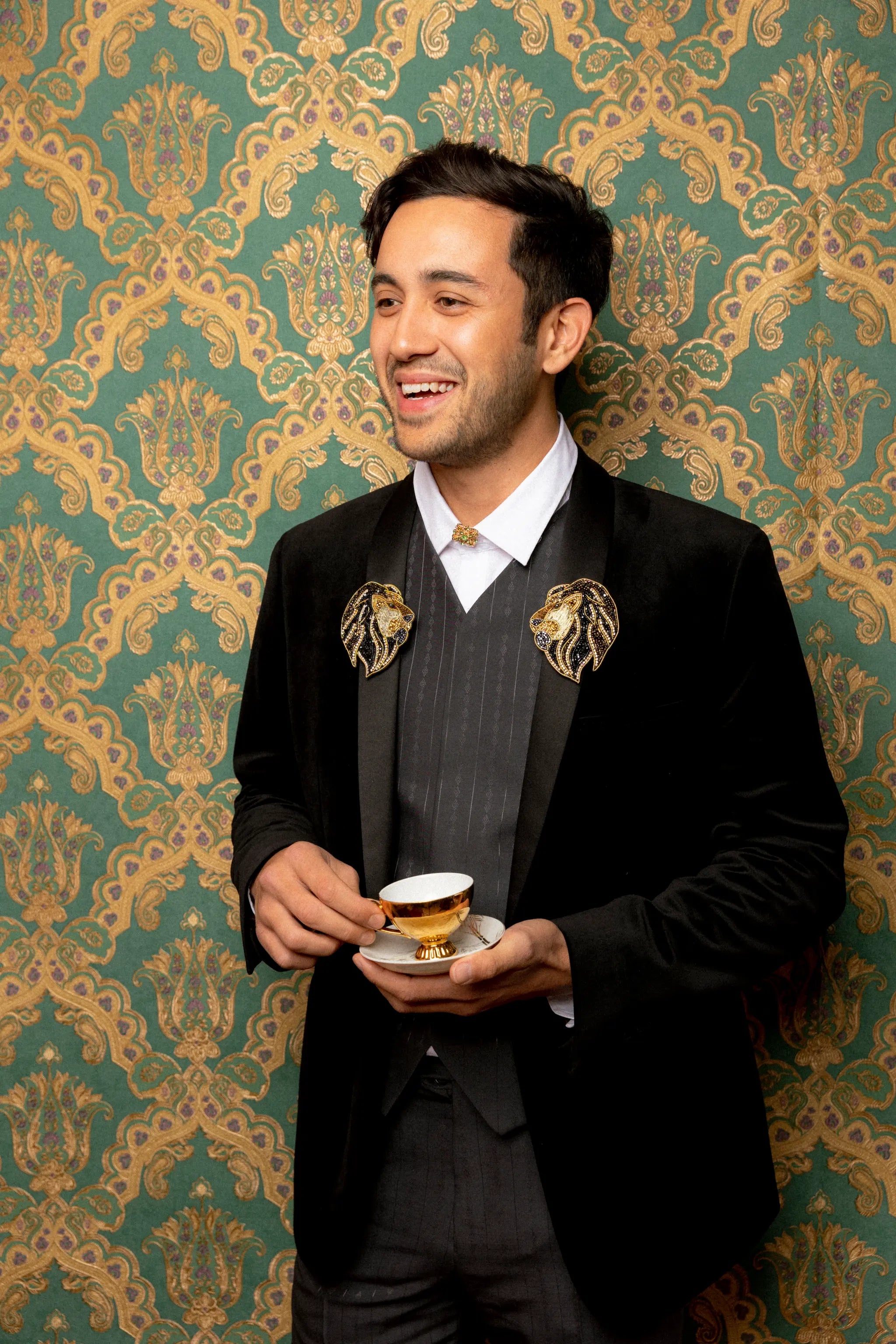 A man in a black velvet tuxedo jacket with Broochella mirrored gold lion brooches on the lapels, a grey pinstripe vest, and a white shirt, holding a teacup and saucer against a green and gold damask wallpaper background.