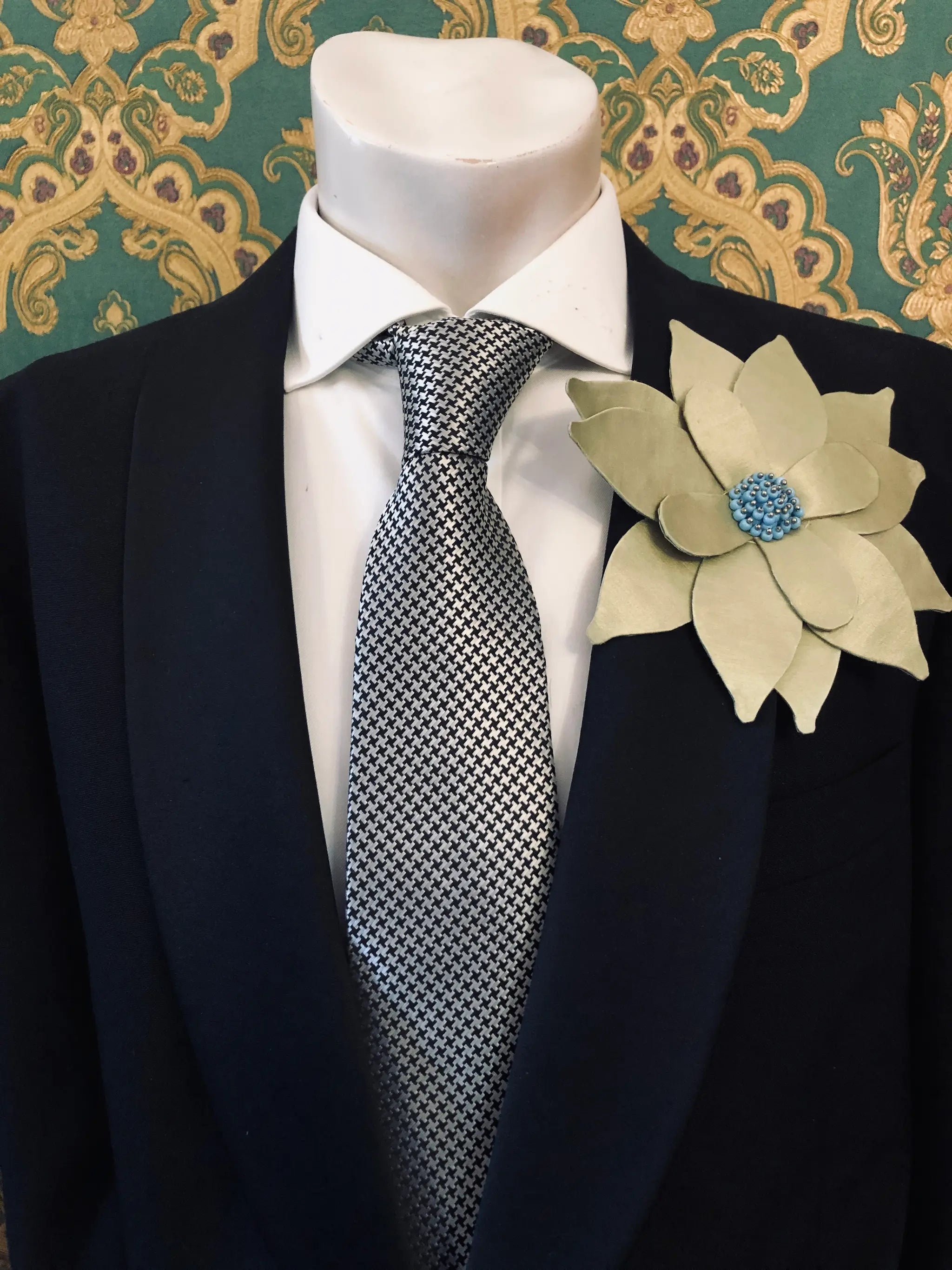 A men's formal wear set displayed on a white mannequin torso, featuring a dark navy suit jacket, a white dress shirt, a silver and black checkered tie, and a large Broochella Men's Ninfea flower brooch with a blue beaded centre on the lapel.