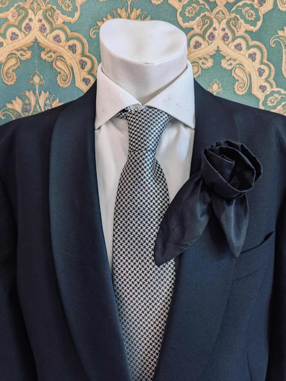 A Broochella Men's Petalo black, small, handmade silk  rose-shaped flower brooch with petals. It is displayed on a men's mannequin that is dress in a black suit, with a silver and black houndstooth tie. It is against a dark teal and gold damask patterned wallpaper background