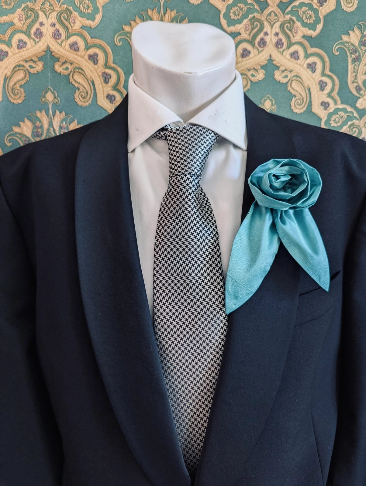 A Broochella Men's Petalo blue, small, handmade silk  rose-shaped flower brooch with petals. It is displayed on a men's mannequin that is dress in a black suit, with a silver and black houndstooth tie. It is against a dark teal and gold damask patterned wallpaper background