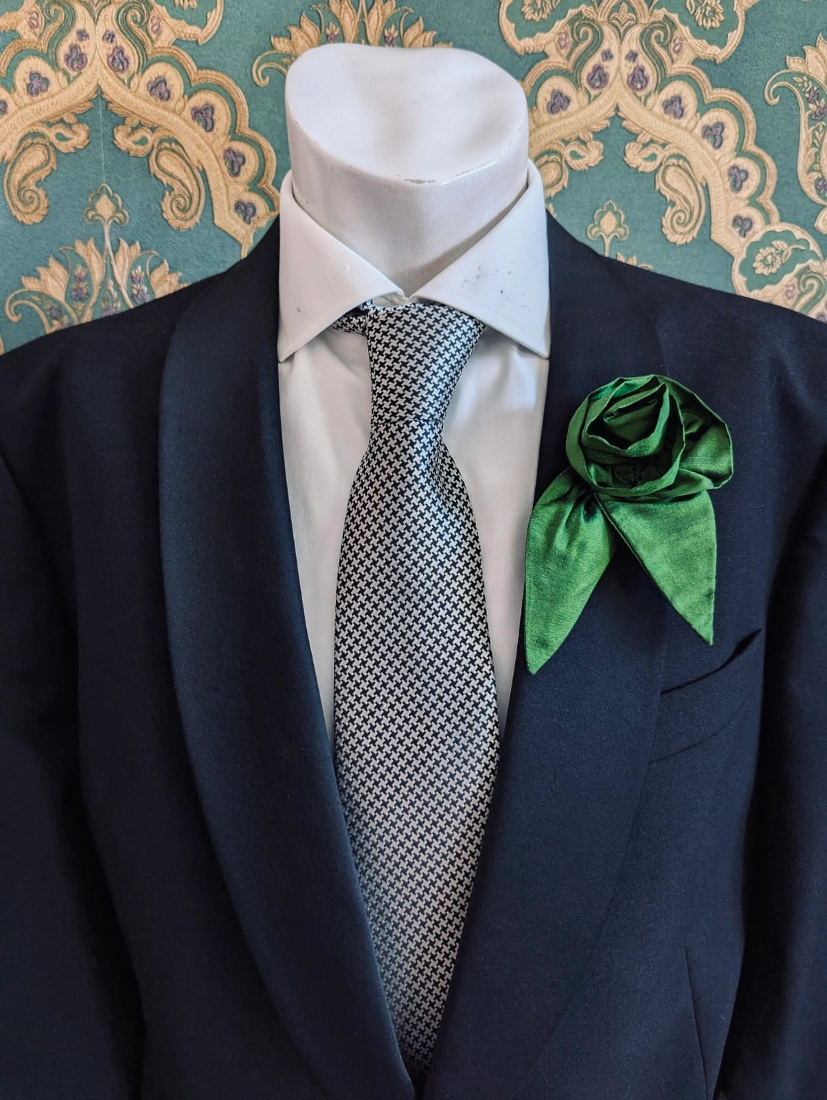 A Broochella Men's Petalo green, small, handmade silk  rose-shaped flower brooch with petals. It is displayed on a men's mannequin that is dress in a black suit, with a silver and black houndstooth tie. It is against a dark teal and gold damask patterned wallpaper background