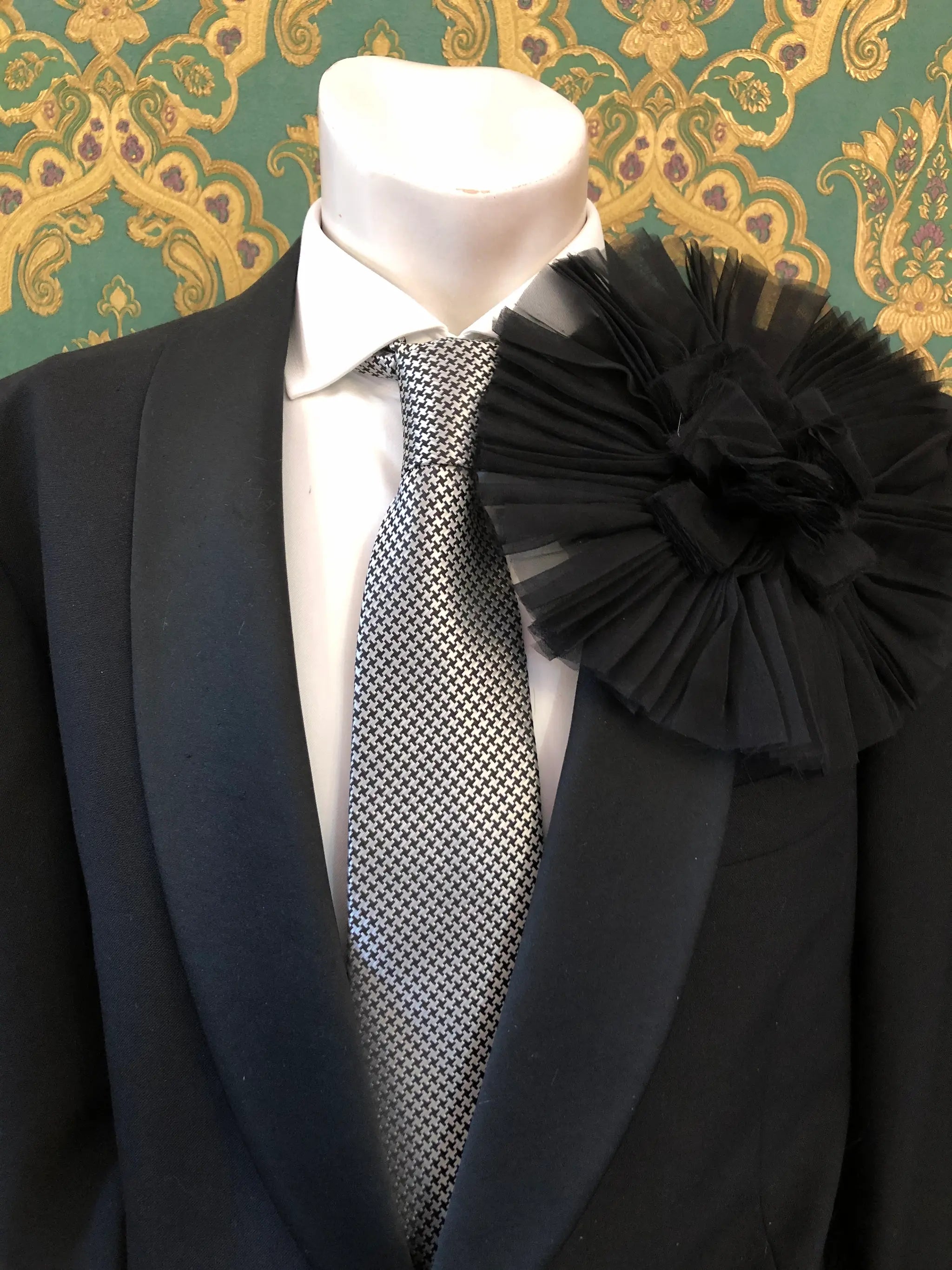 A formal outfit displayed on a mannequin torso, featuring a black suit jacket, white dress shirt, patterned grey and white tie, and a large Broochella Men's Pieghettato black flower brooch on the lapel. The background is ornate green and gold damask wallpaper.