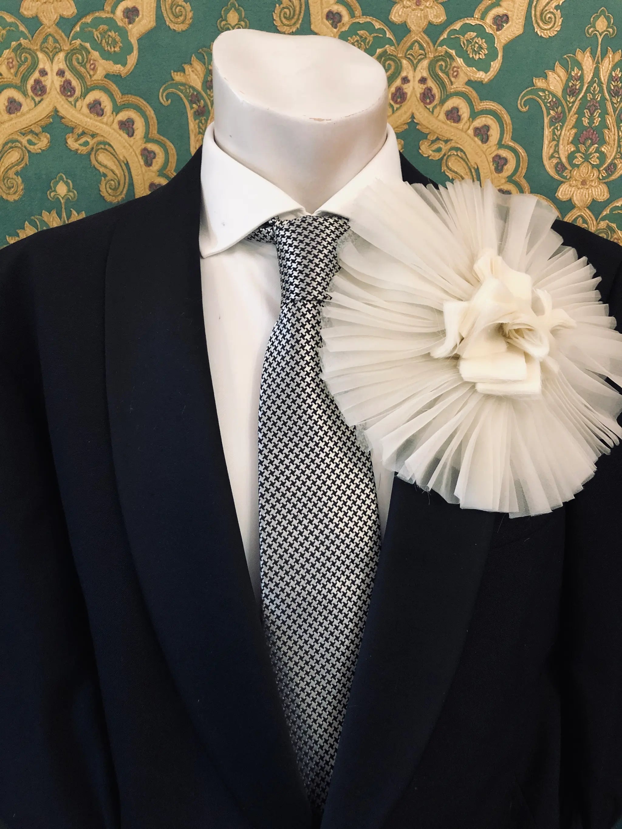 A formal outfit displayed on a mannequin torso, featuring a black suit jacket, white dress shirt, patterned grey and white tie, and a large Broochella Men's Pieghettato ivory flower brooch on the lapel. The background is ornate green and gold damask wallpaper.