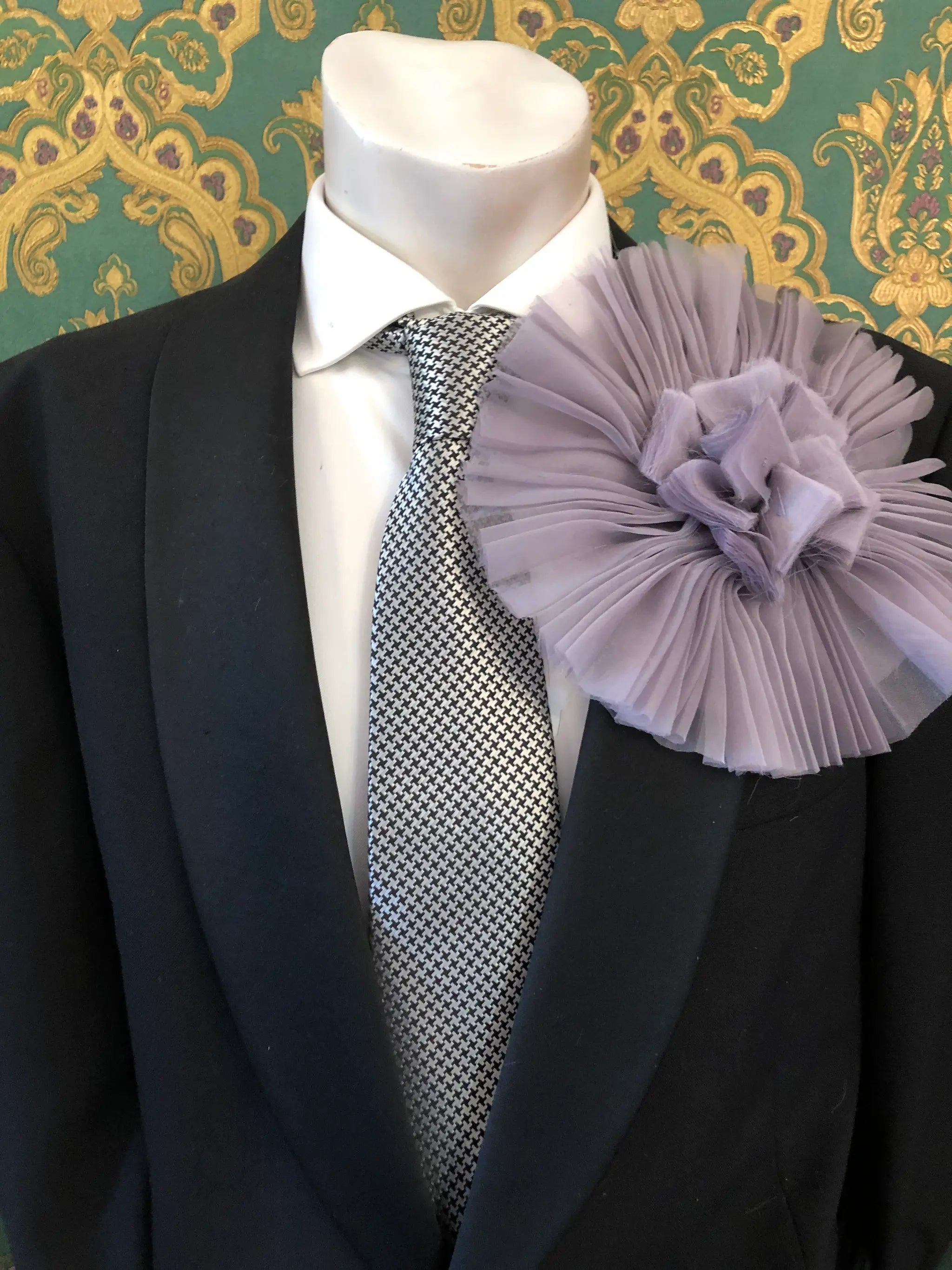 A formal outfit displayed on a mannequin torso, featuring a black suit jacket, white dress shirt, patterned grey and white tie, and a large Broochella Men's Pieghettato lavender flower brooch on the lapel. The background is ornate green and gold damask wallpaper.