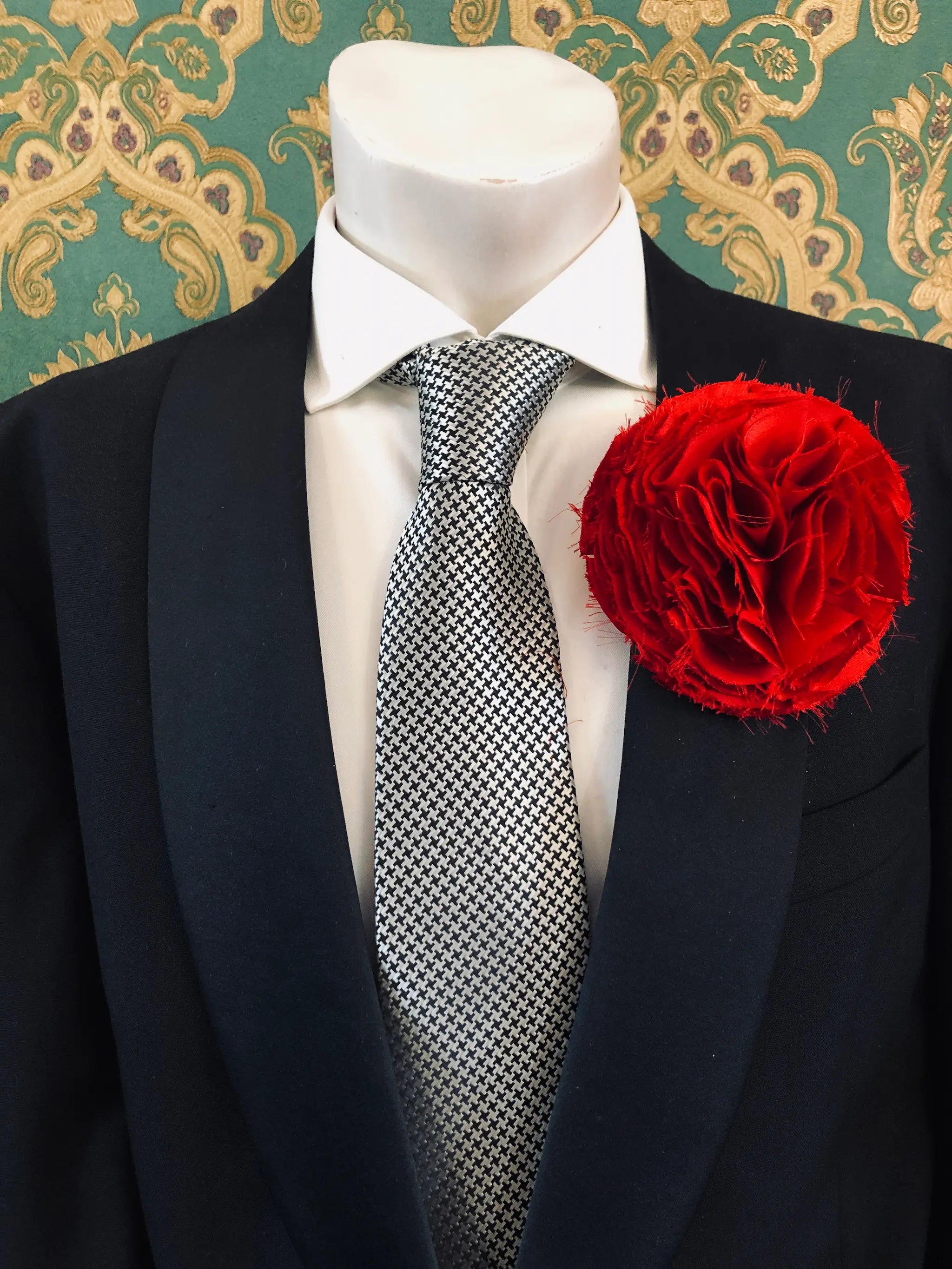 A men's formal wear ensemble displayed on a white mannequin torso, featuring a black suit jacket, a white dress shirt, a black and white checkered pattern tie, and a vibrant Broochella Men's Pom Pon green textured fabric flower brooch.