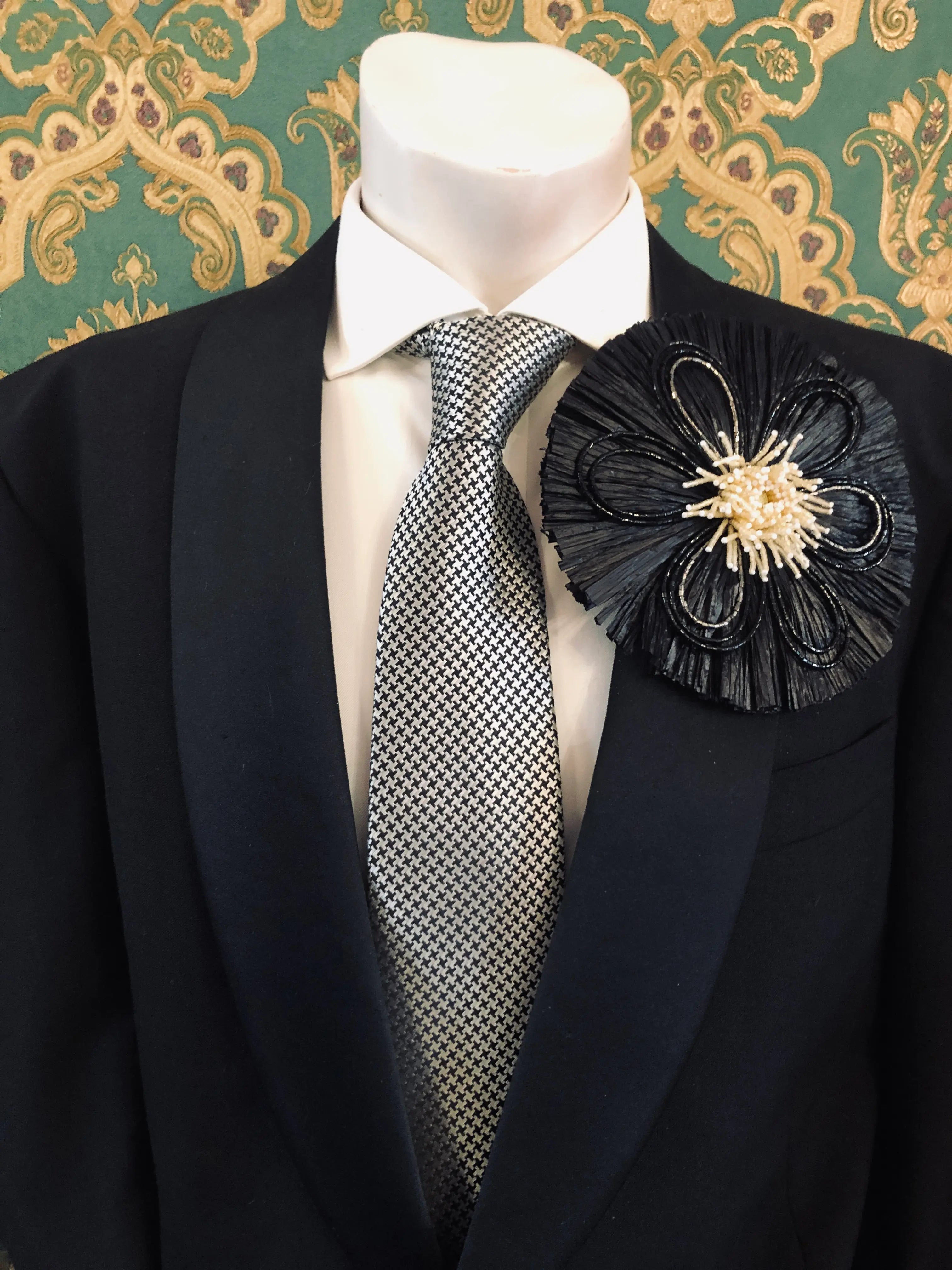 A Broochella Men's Raffia Bloom black fabric flower-shaped brooch pin with a gold and white beaded centre, displayed on a black suit jacket with a patterned grey tie and white shirt, set against a green and gold patterned wallpaper background.