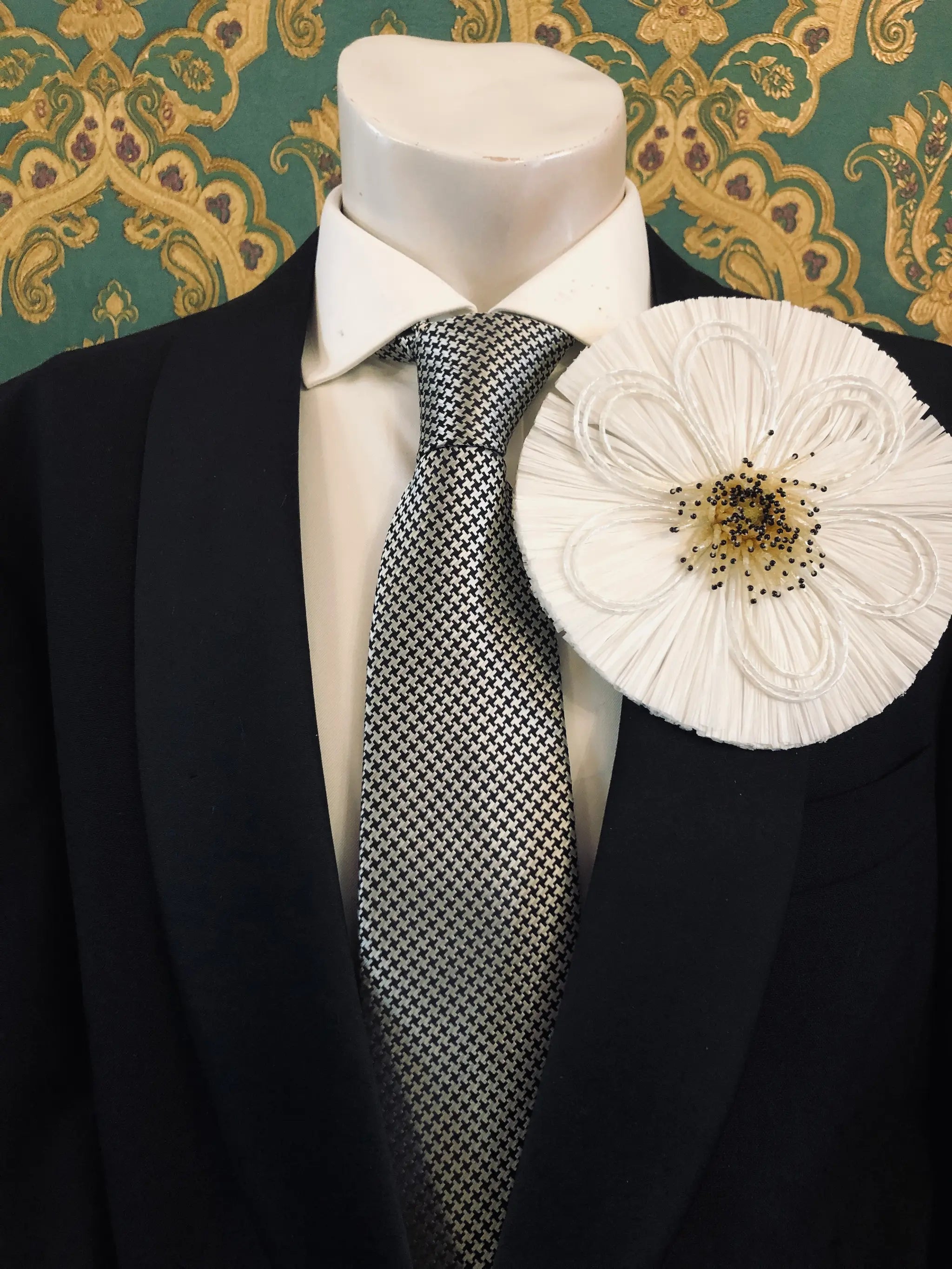 A Broochella Men's Raffia Bloom black fabric flower-shaped brooch pin with a gold and white beaded centre, displayed on a black suit jacket with a patterned grey tie and white shirt, set against a green and gold patterned wallpaper background.