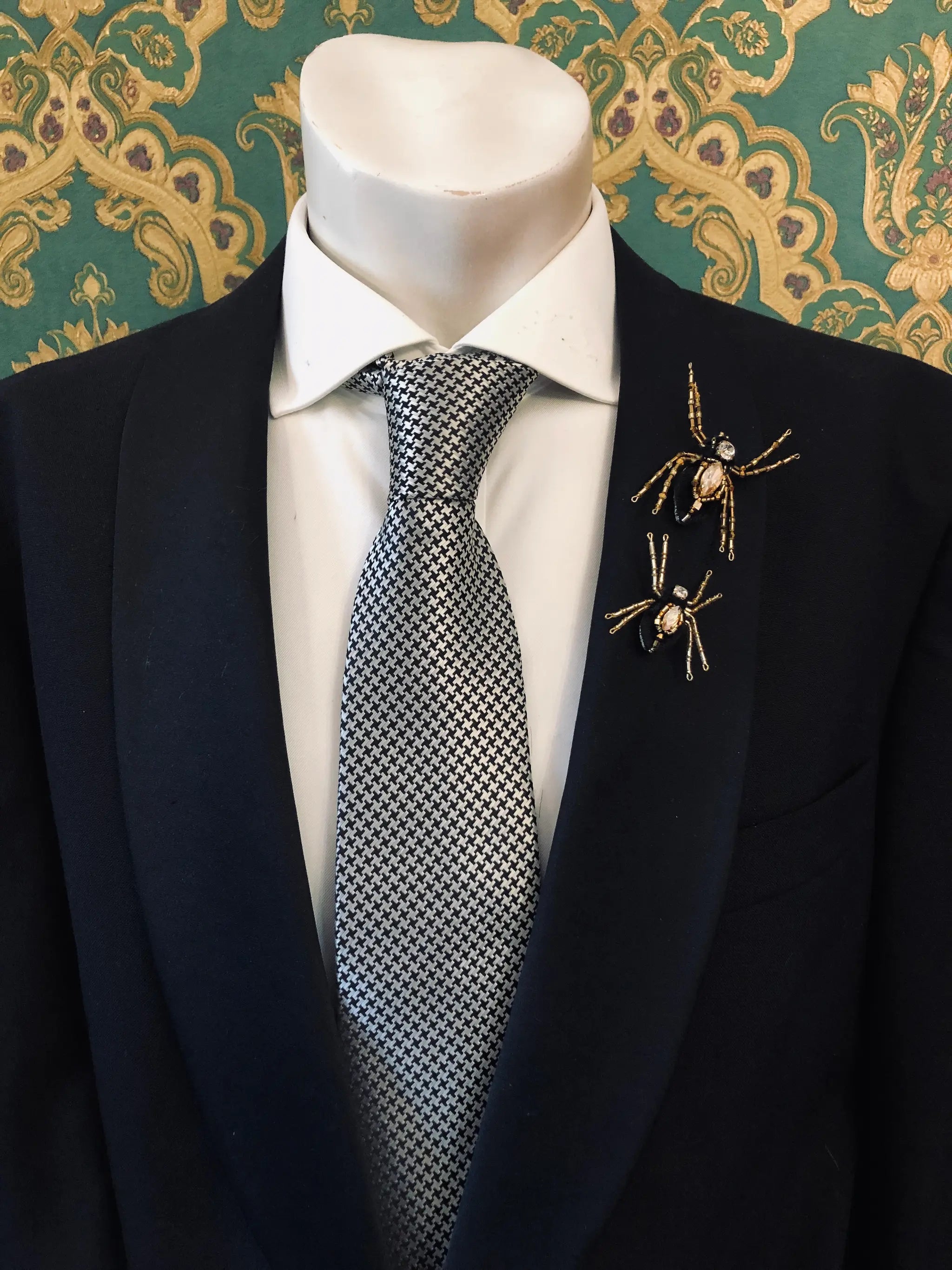 A mannequin torso wearing a black suit jacket, white dress shirt, and a grey-and-white patterned tie, with two Broochella men's gold spider-shaped lapel pins on the left lapel. The background is ornate green and gold patterned wallpaper.