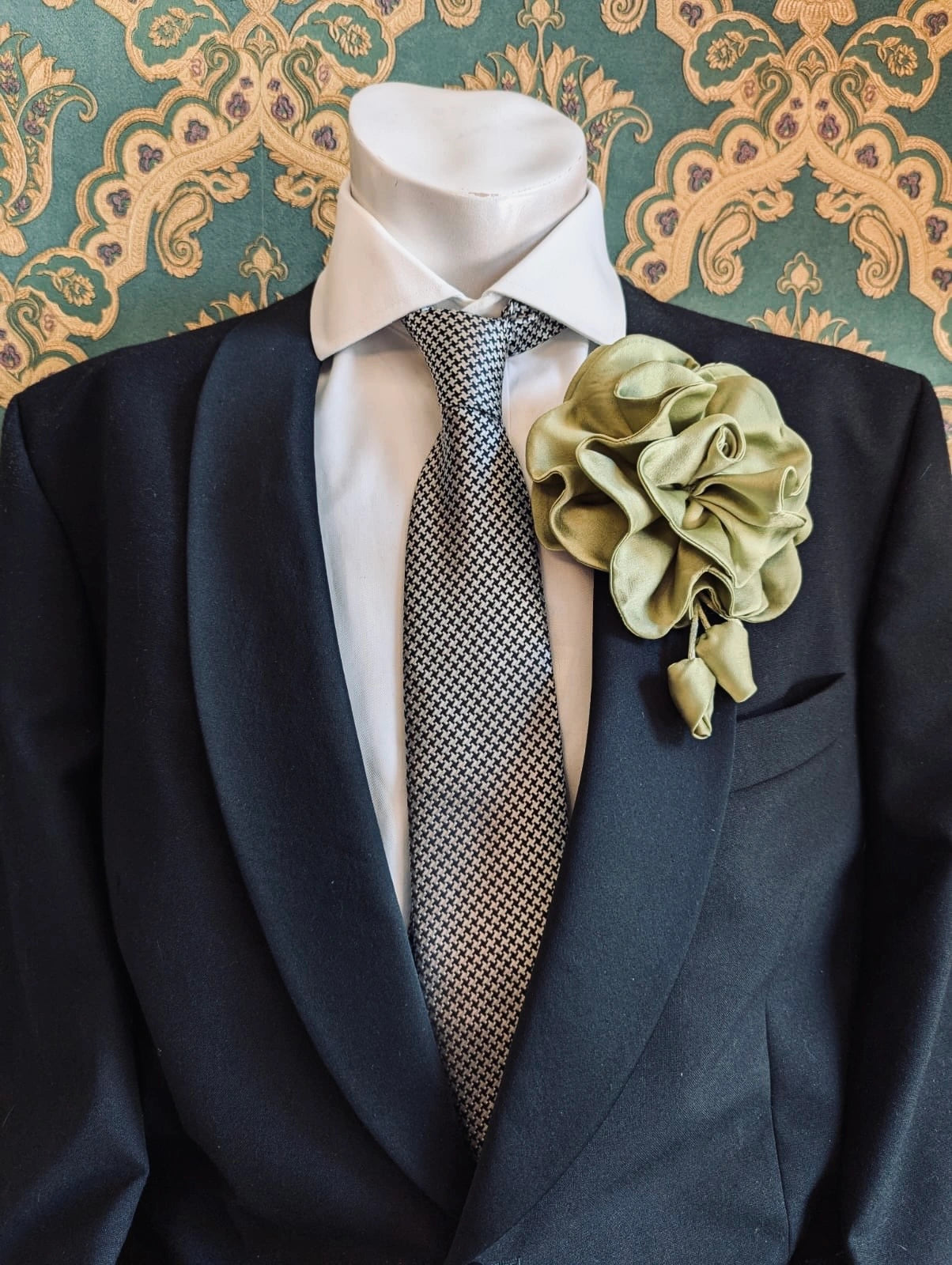 Broochella men's rosetta handmade, designer, satin flower brooch in avocado. Pinned on a men's mannequin wearing a black suit and a houndstooth tie. The background is green and gold damask wallpaper. 