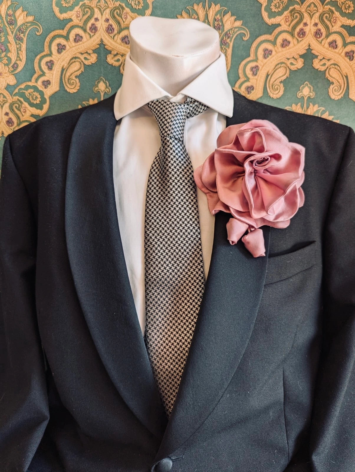 Broochella men's rosetta handmade, designer, satin flower brooch in pink. Pinned on a men's mannequin wearing a black suit and a houndstooth tie. The background is green and gold damask wallpaper. 
