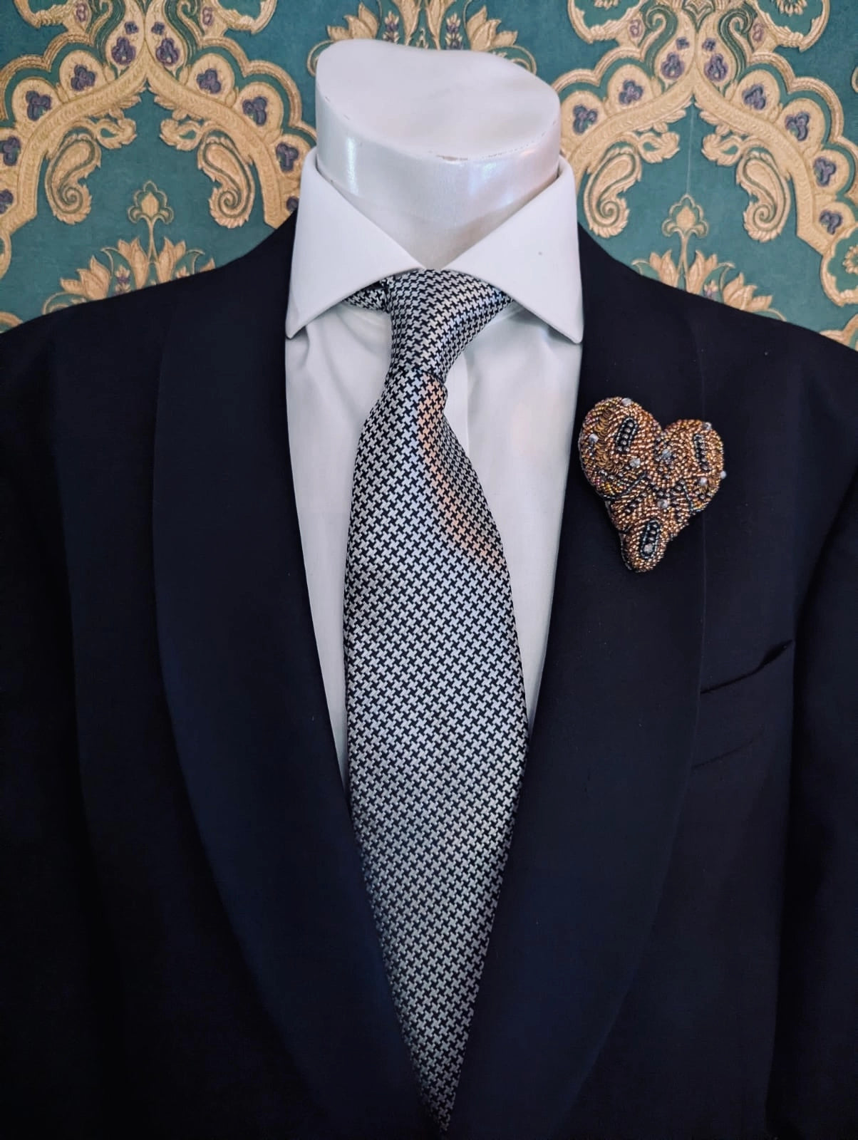 Broochella men's amore hand beaded heart shaped brooch in bronze and silver beads and sequins. Pinned to a men's mannequin wearing a black suit and a tie. The background is green and gold damask wallpaper.