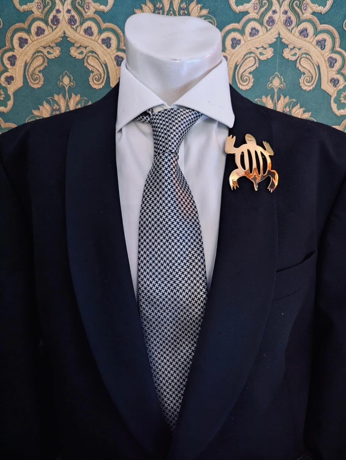 Broochella men's brooches with meaning adaptability gold metal turtle brooch. Pinned to a men's mannequin wearing a black suit and houndstooth tie. The background is green and gold damask wallpaper.