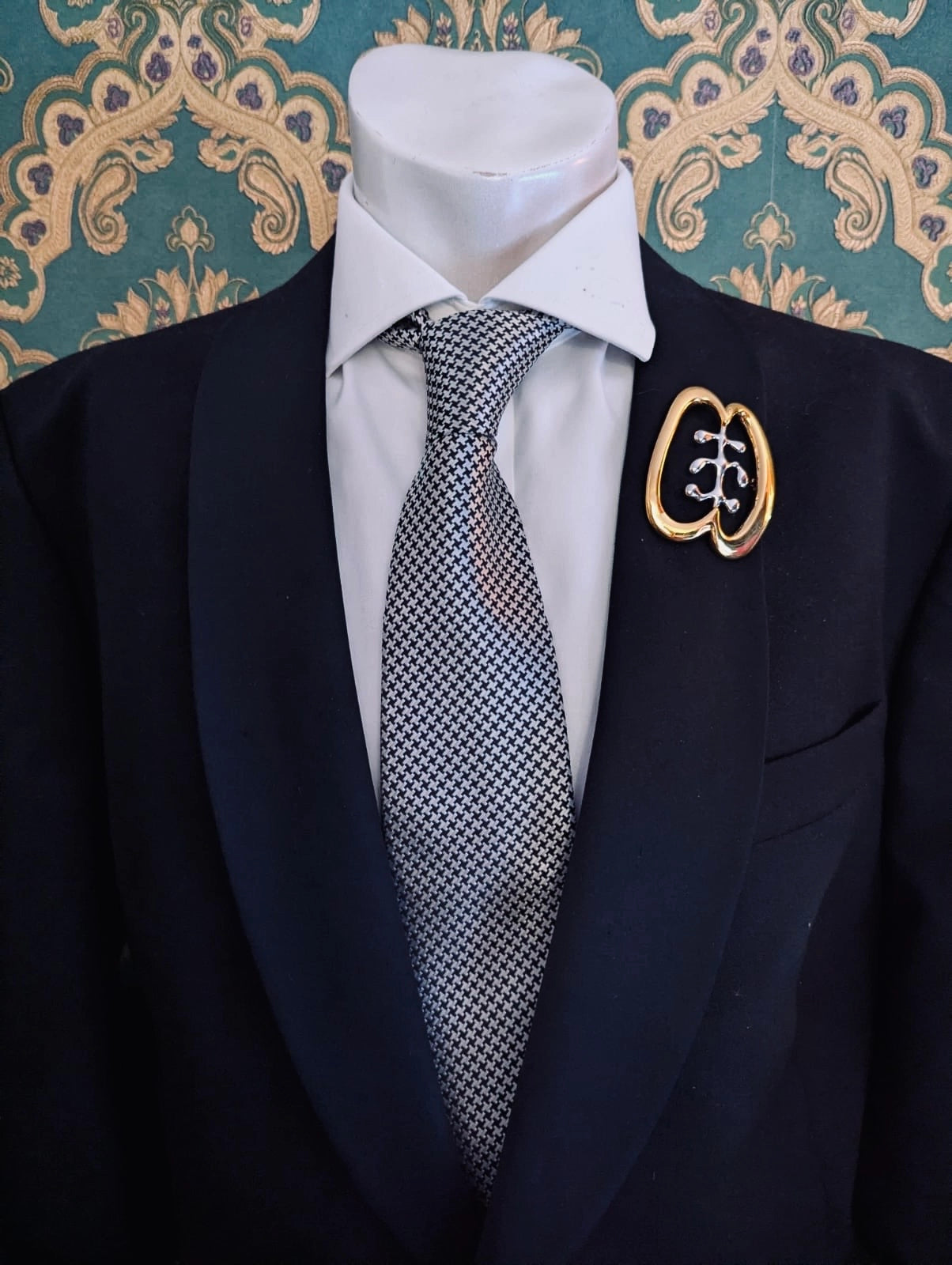 Broochella men's brooches with meaning friendship gold metal and silver brooch. Pinned to a men's mannequin wearing a black suit and houndstooth tie. The background is green and gold damask wallpaper.