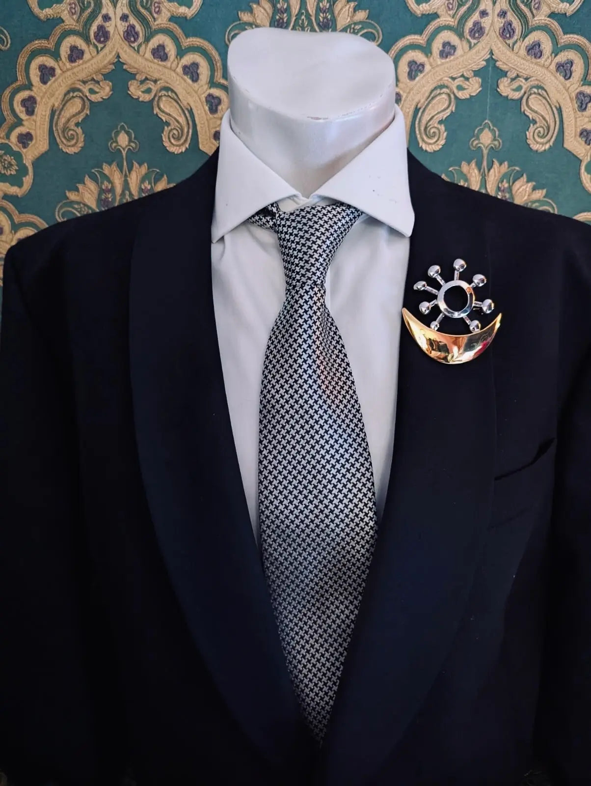 Broochella men's brooches with meaning love and harmony gold and silver metal brooch. Pinned to a men's mannequin wearing a black suit and houndstooth tie. The background is green and gold damask wallpaper.