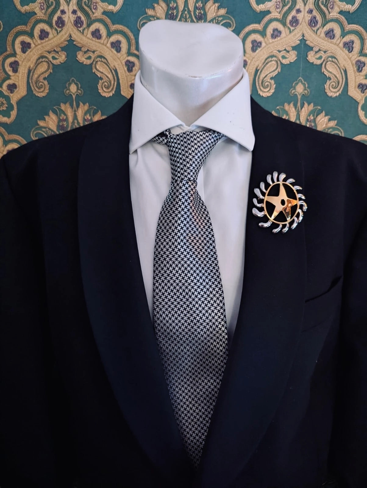 Broochella men's brooches with meaning welcoming change gold metal and silver brooch. Pinned to a men's mannequin wearing a black suit and houndstooth tie. The background is green and gold damask wallpaper.