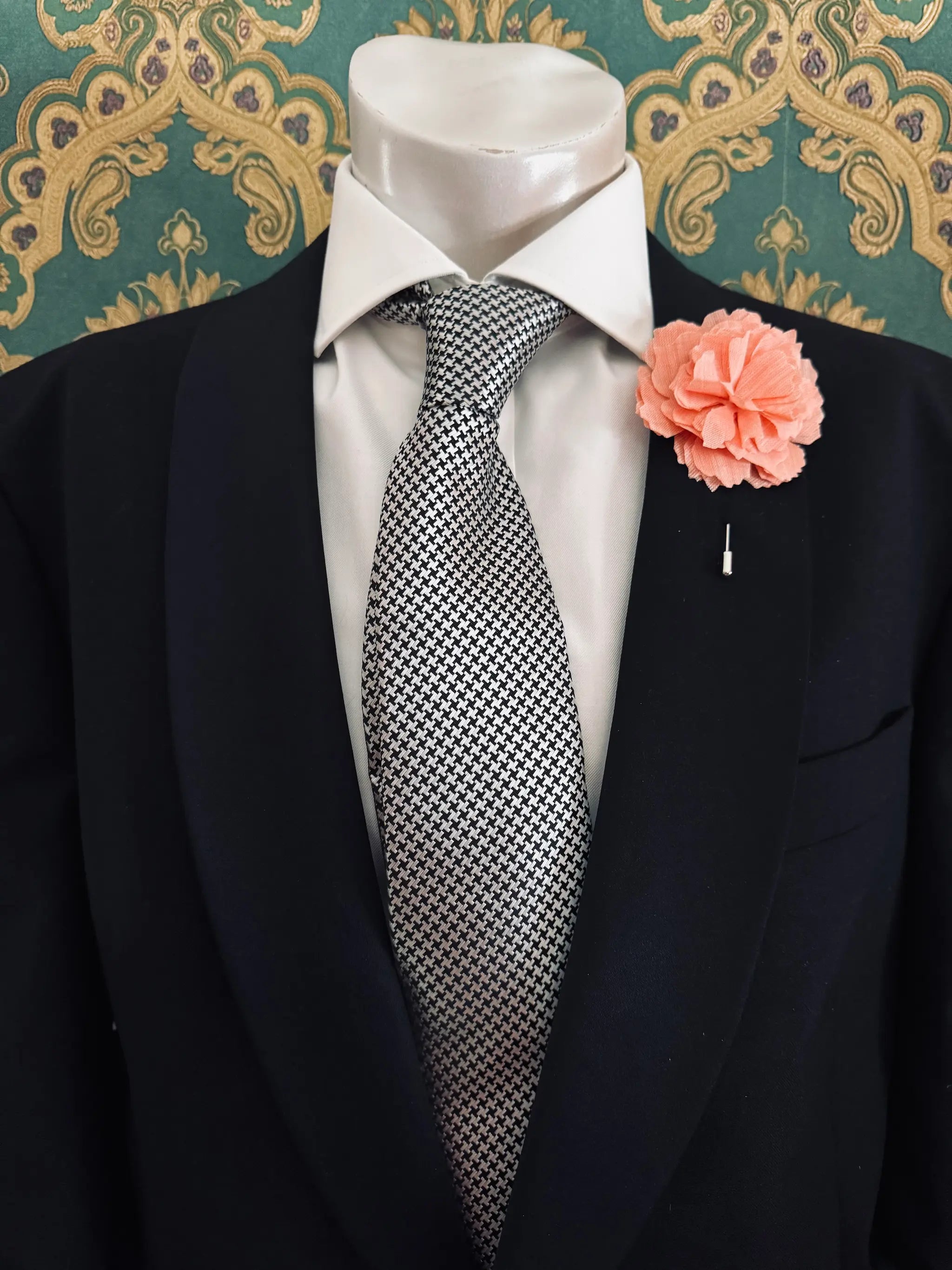 Broochella men's handmade linen carnation brooch in orange. Pinned to a men's mannequin wearing a black suit and a tie. The background is green and gold damask wallpaper.
