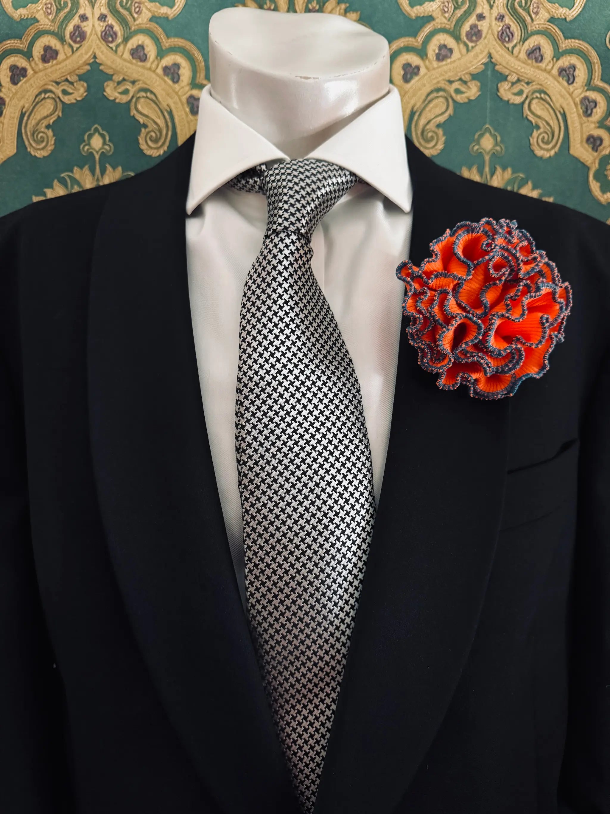 Broochella Men's corallo brooch. Made from orange, ribbed, stretch fabric. The edges are finished with blue stitching.  The brooch resembles a bright piece of coral. Pinned to a men's mannequin wearing a black suit and a tie. The background is green and gold damask wallpaper.