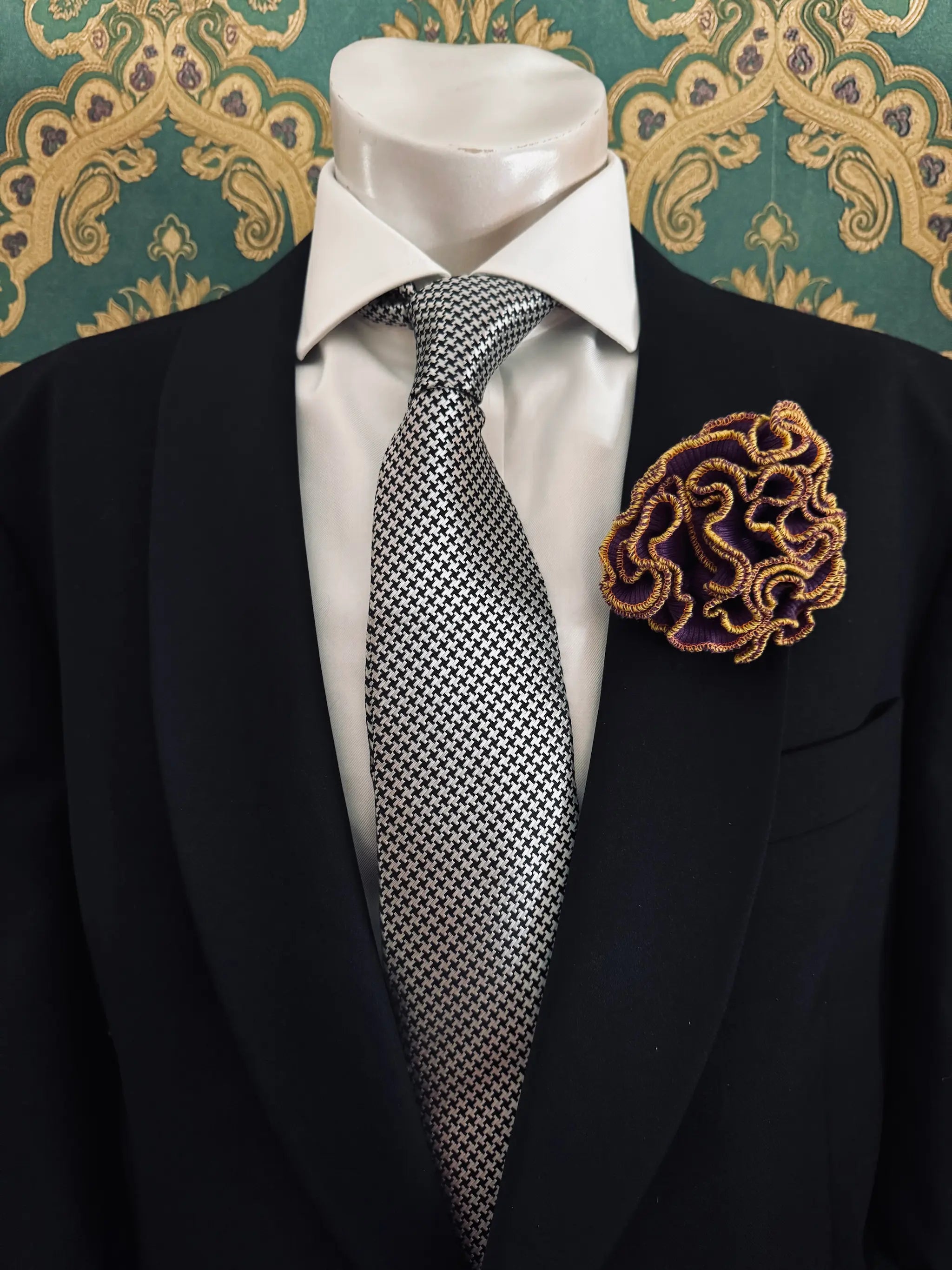 Broochella Men's corallo brooch. Made from purple, ribbed, stretch fabric. The edges are finished with yellow stitching.  The brooch resembles a bright piece of coral. Pinned to a men's mannequin wearing a black suit and a tie. The background is green and gold damask wallpaper.