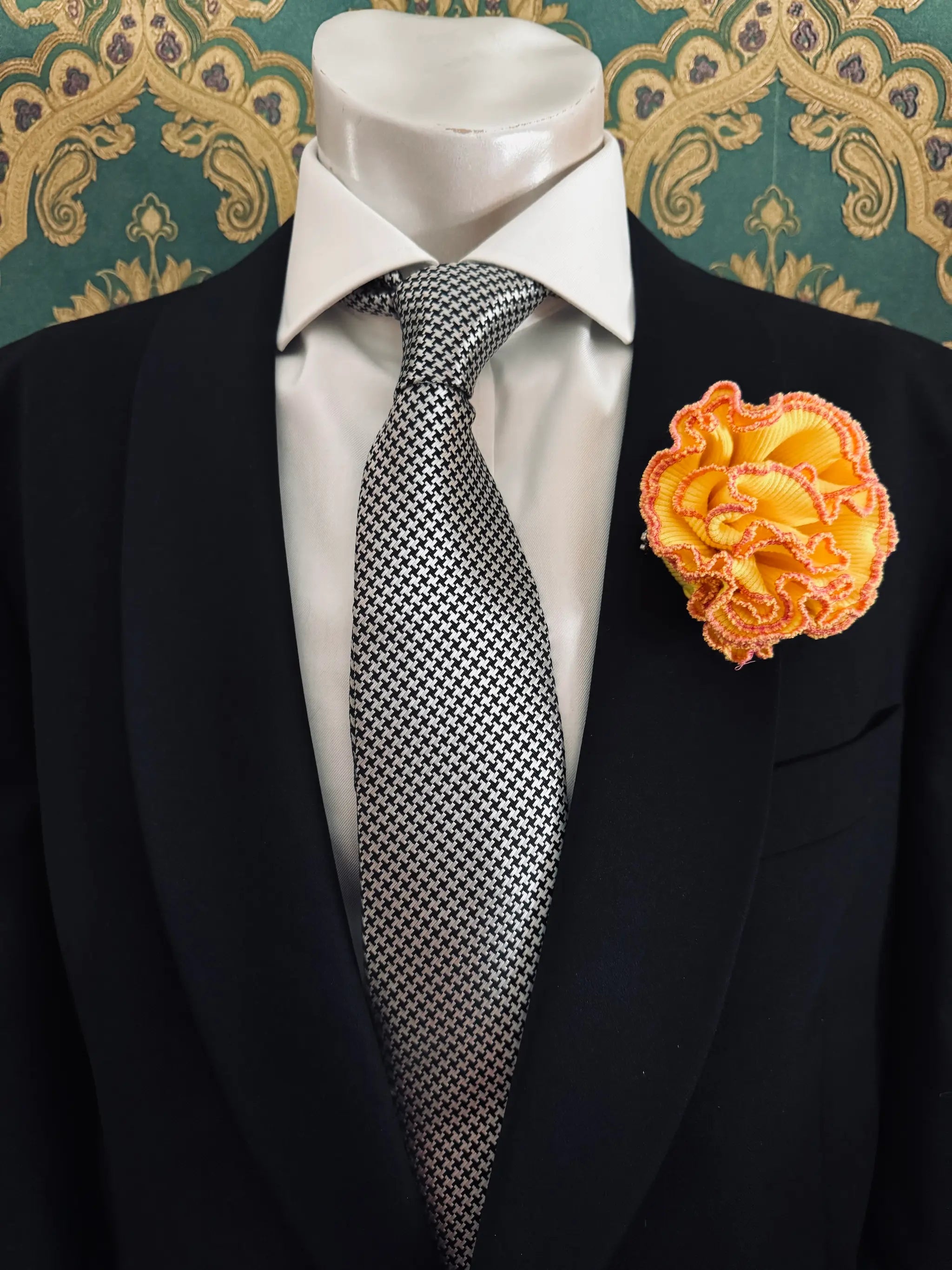 Broochella Men's corallo brooch. Made from yellow, ribbed, stretch fabric. The edges are finished with pink stitching.  The brooch resembles a bright piece of coral. Pinned to a men's mannequin wearing a black suit and a tie. The background is green and gold damask wallpaper.