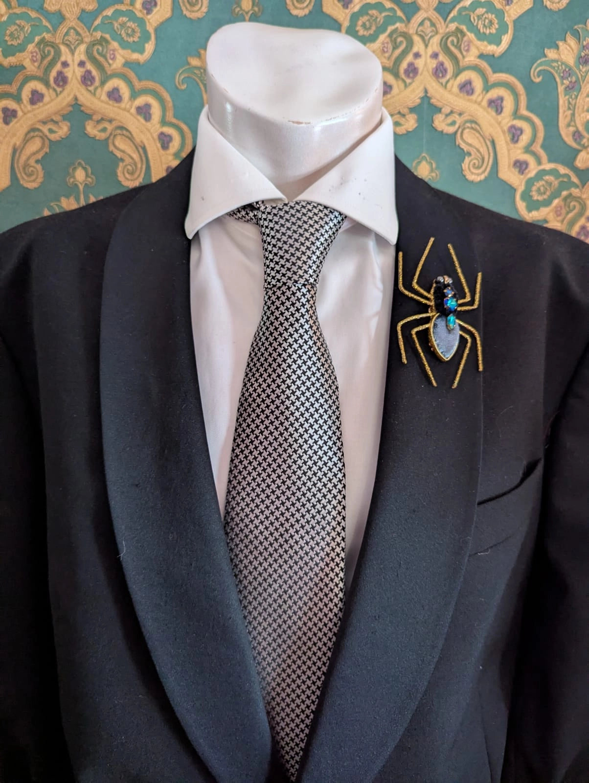 Broochella  femme fatale hand beaded gold and blue spider brooch. The brooch is pinned on a men's mannequin in a black suit The background is green and gold damask wallpaper.