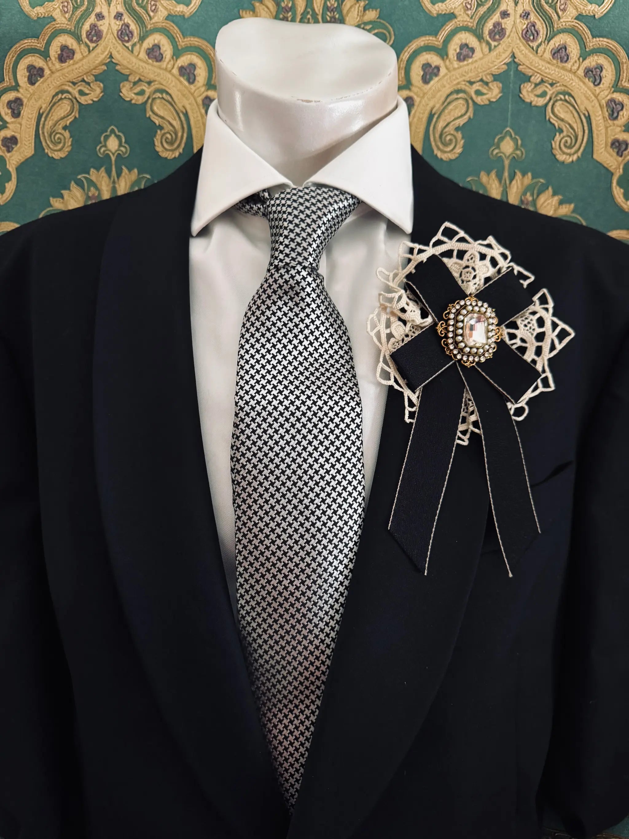 Broochella men's handmade il conte black bow brooch with crochet lace backing and a crystal embellishment centre. Pinned to a men's mannequin wearing a black suit and a tie. The background is green and gold damask wallpaper.