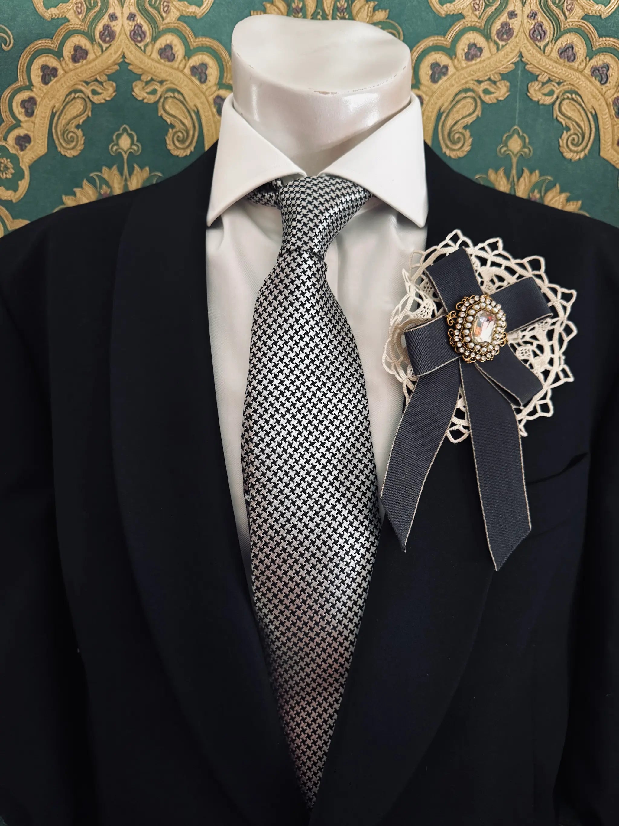 Broochella men's handmade il conte grey bow brooch with crochet lace backing and a crystal embellishment centre. Pinned to a men's mannequin wearing a black suit and a tie. The background is green and gold damask wallpaper.