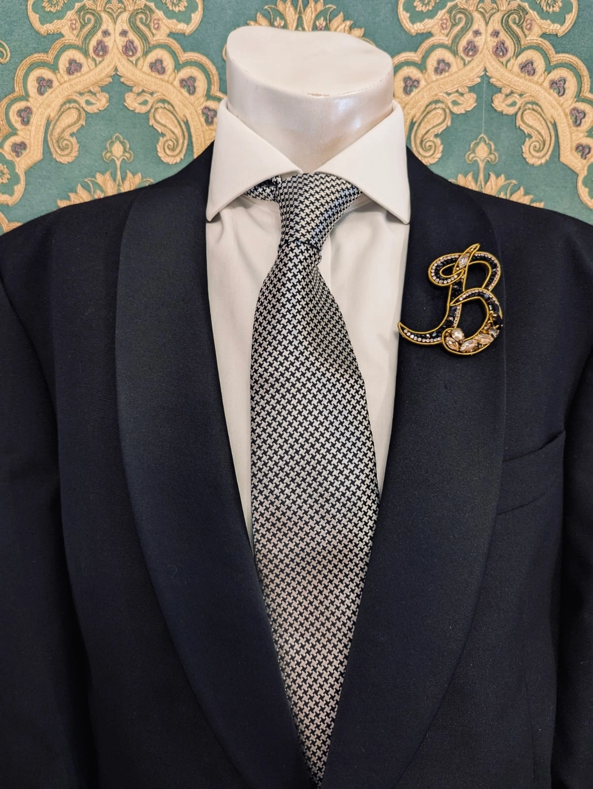 Broochella men's hand beaded B initial brooch in black and gold beads and sequins. Pinned to a men's mannequin wearing a black suit and a tie. The background is green and gold damask wallpaper.